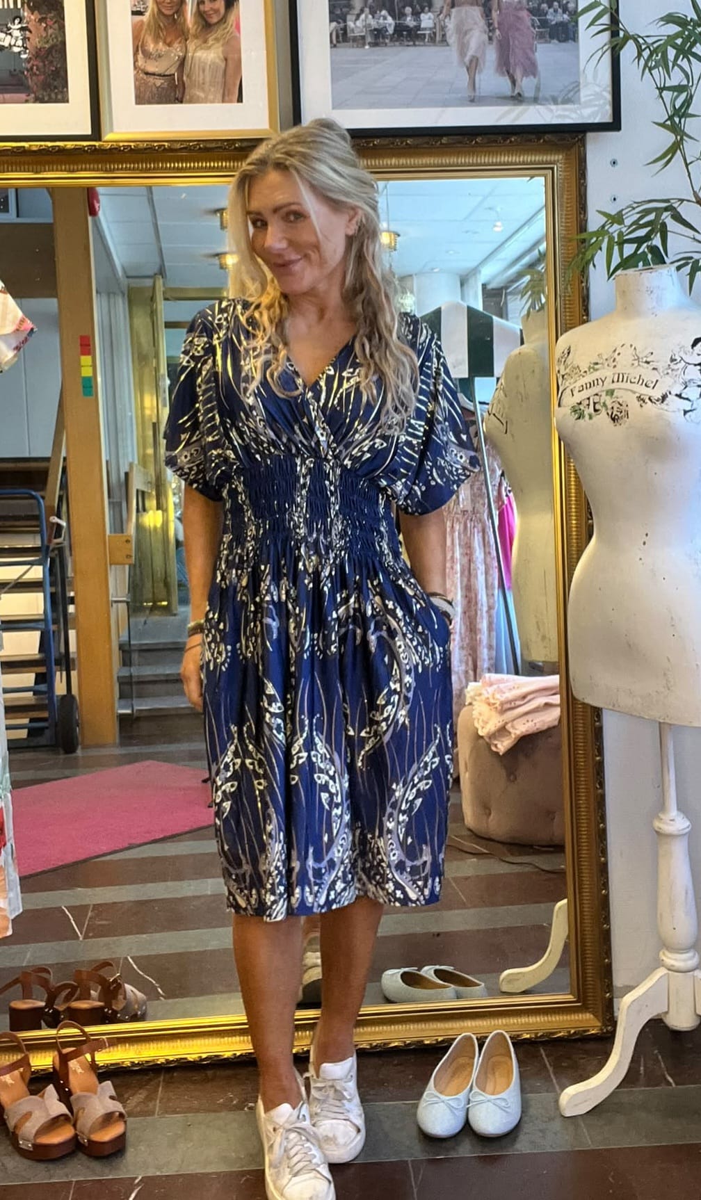 Kvinna står och ler framför en stor guldramsspegel, iklädd blå mönstrad klänning och vita sneakers. Runt henne syns en provdocka med texten “Fanny Michel” och flera par skor i en butiksmiljö.