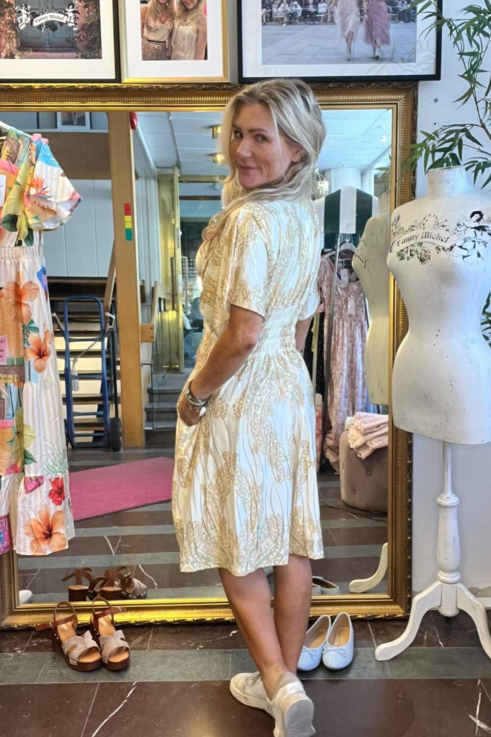 Kvinna provar en vit klänning med guldmönster och vänder sig leende mot kameran i en klädbutik, framför en stor spegel. Runt henne syns skor på golvet, provdocka och inramade foton; text: “CASA FANNY”, “Fanny Michel”.