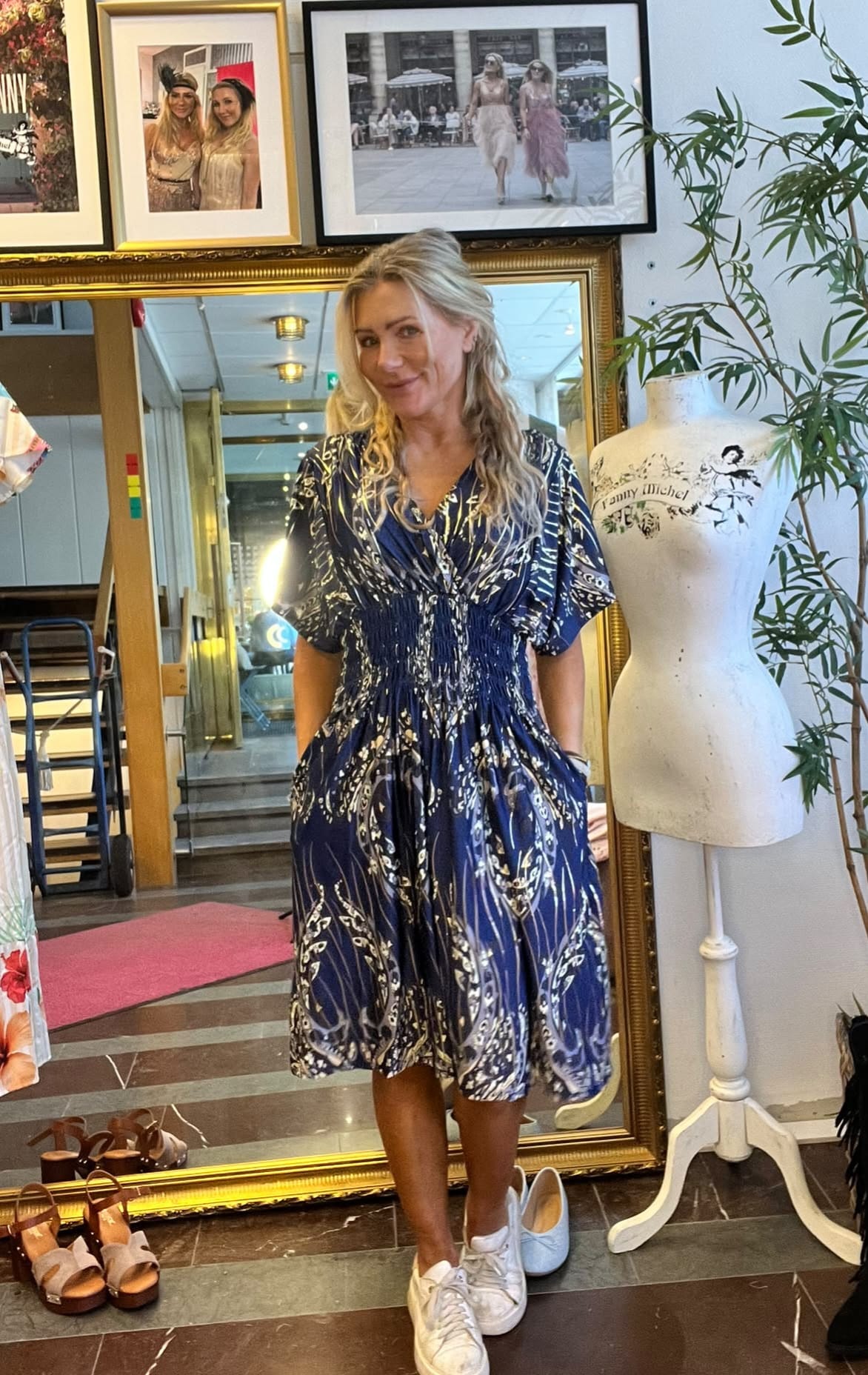 Kvinna poserar med händerna i fickorna och ler, iklädd blåmönstrad klänning och vita sneakers, framför en stor guldramsspegel i en butik. Bredvid står en provdocka med texten ”Ronny michel”, och inramade foton hänger ovanför.