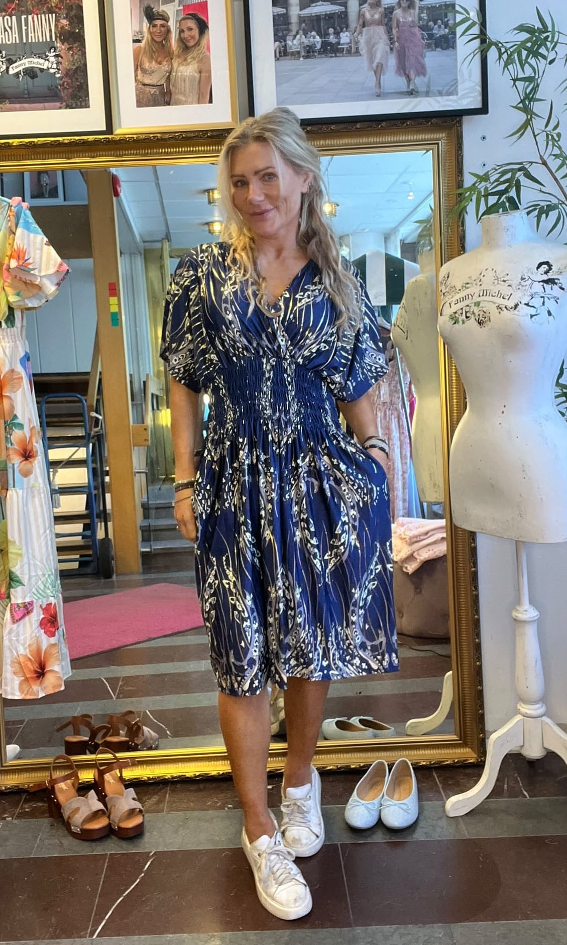 Blond kvinna står och ler i en klädbutik, med handen i fickan på en blå, mönstrad klänning och vita sneakers. Bakom henne syns en stor guldramad spegel, skor på golvet och en provdocka med texten “Fanny Michel”.
