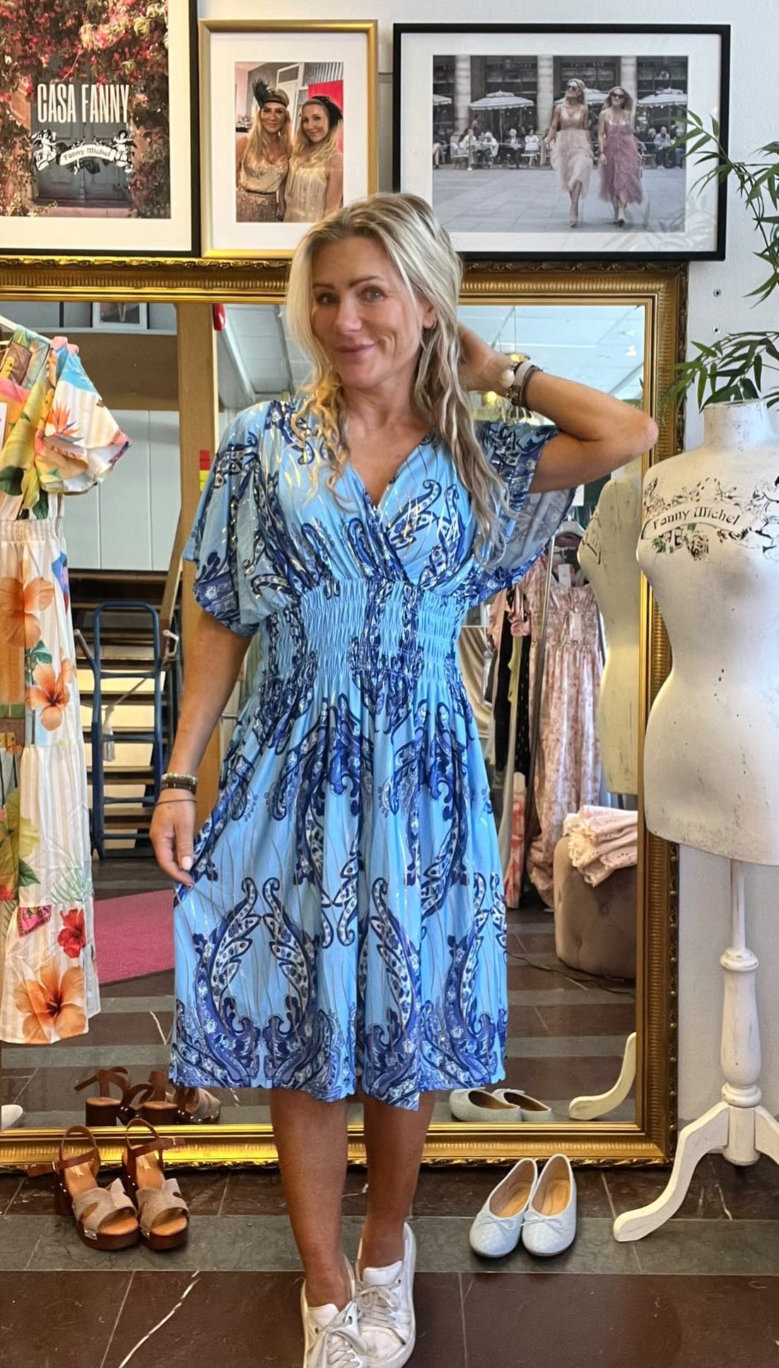 Kvinna poserar i en klädbutik framför en stor guldspegel, håller ena handen i håret och visar en blå mönstrad klänning och vita sneakers. Runtom syns skor, klänningar, provdocka och inramade foton med texten ”CASA FANNY”.