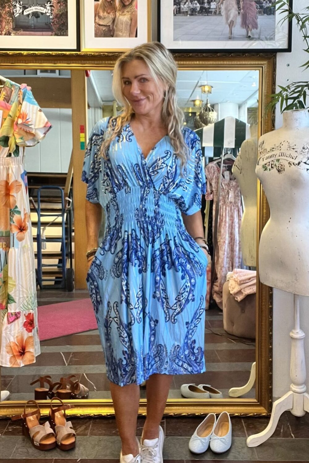 Blond kvinna står framför en stor guldspegel i en klädbutik och ler med händerna i fickorna. Hon bär en ljusblå, mönstrad klänning och vita sneakers. Runt henne syns skor, klänningar, skyltdocka och tavlor med texten “CASA FANNY” och “Fanny Michel”.