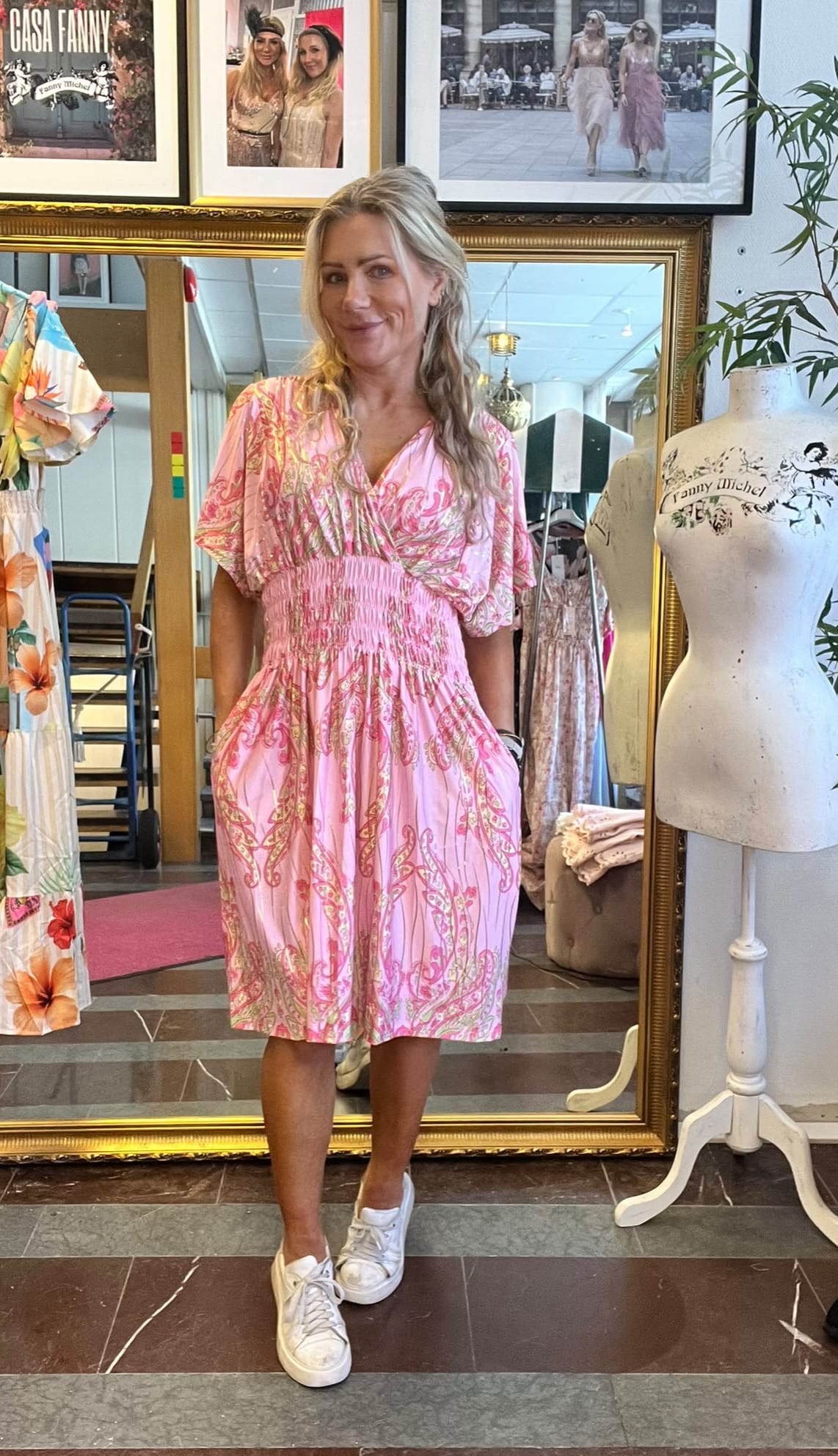 Blond kvinna poserar framför en stor guldramsspegel och provar en rosa mönstrad klänning med smockad midja och vita sneakers. I bakgrunden syns kläder, en provdocka med texten “Fanny Michel” och inramade foton.