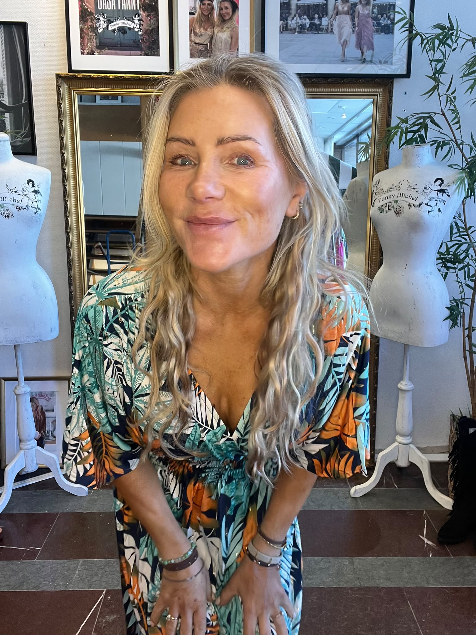 Blond kvinna lutar sig fram och ler mot kameran, klädd i en färgstark klänning med bladmotiv. Hon står mellan två provdockor med texten ”Fanny Michel”, med inramade foton och en spegel på väggen bakom.