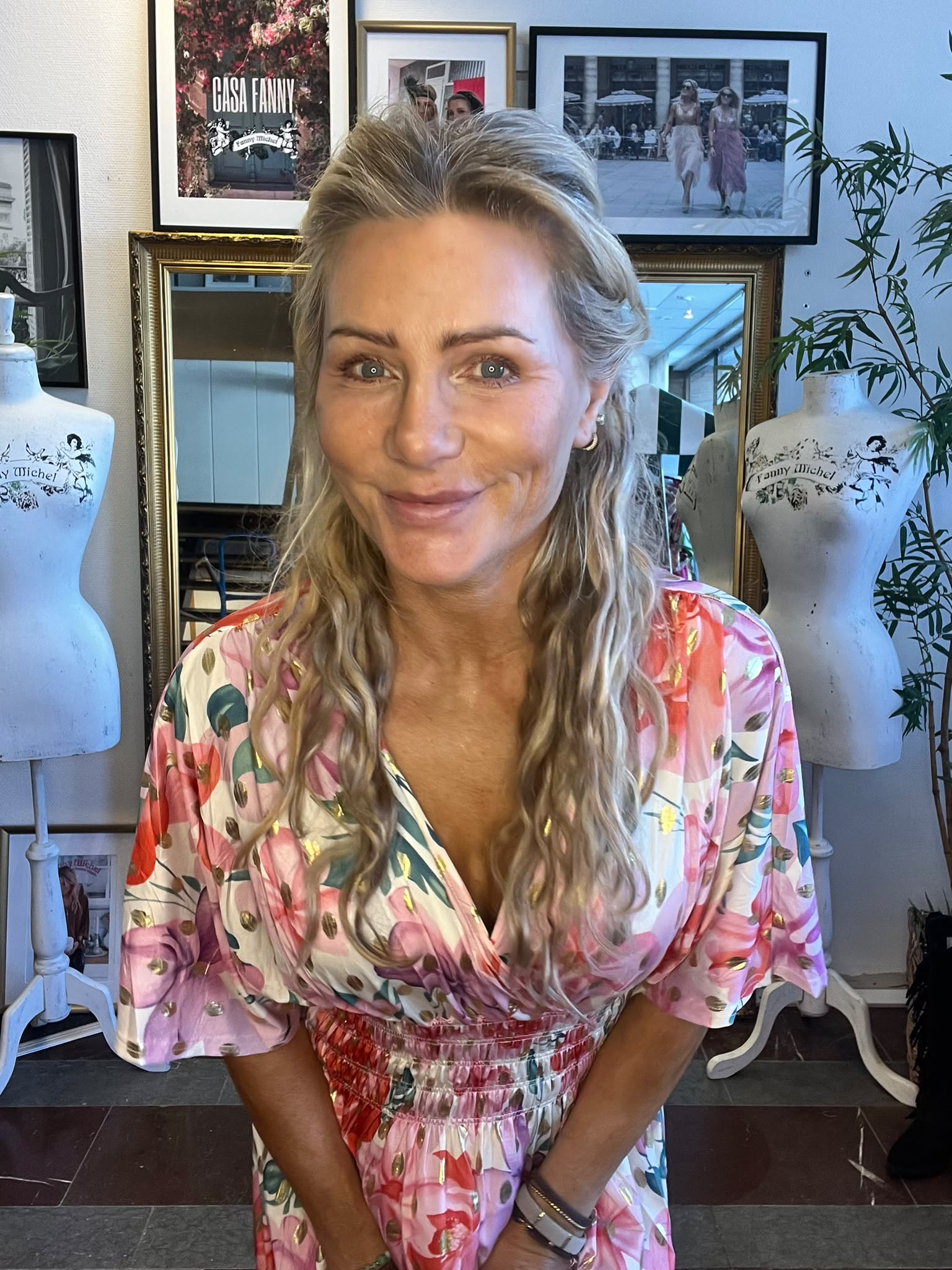 Leende kvinna i en rosa, mönstrad klänning lutar sig mot kameran i en studio. Bakom syns spegel, provdockor med texten “Fanny Michel”, växt och inramade foton, inklusive skylten “CASA FANNY”.