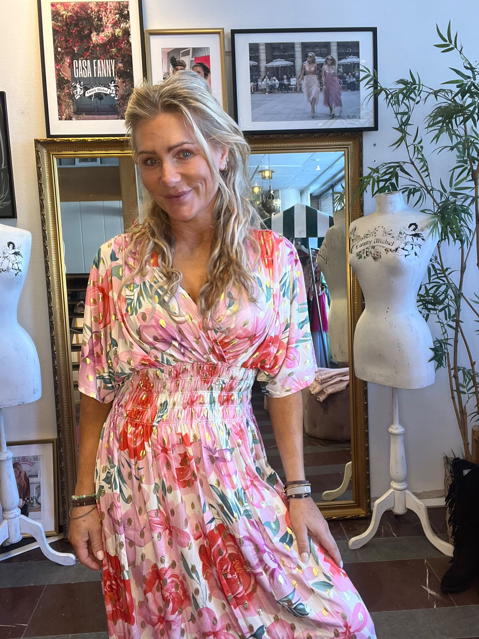 Leende kvinna i rosa blommig klänning poserar inomhus och håller ut kjolen lätt. Bakom henne syns en stor spegel, skyltdockor med texten “Fanny Michel” samt inramade foton och växt.
