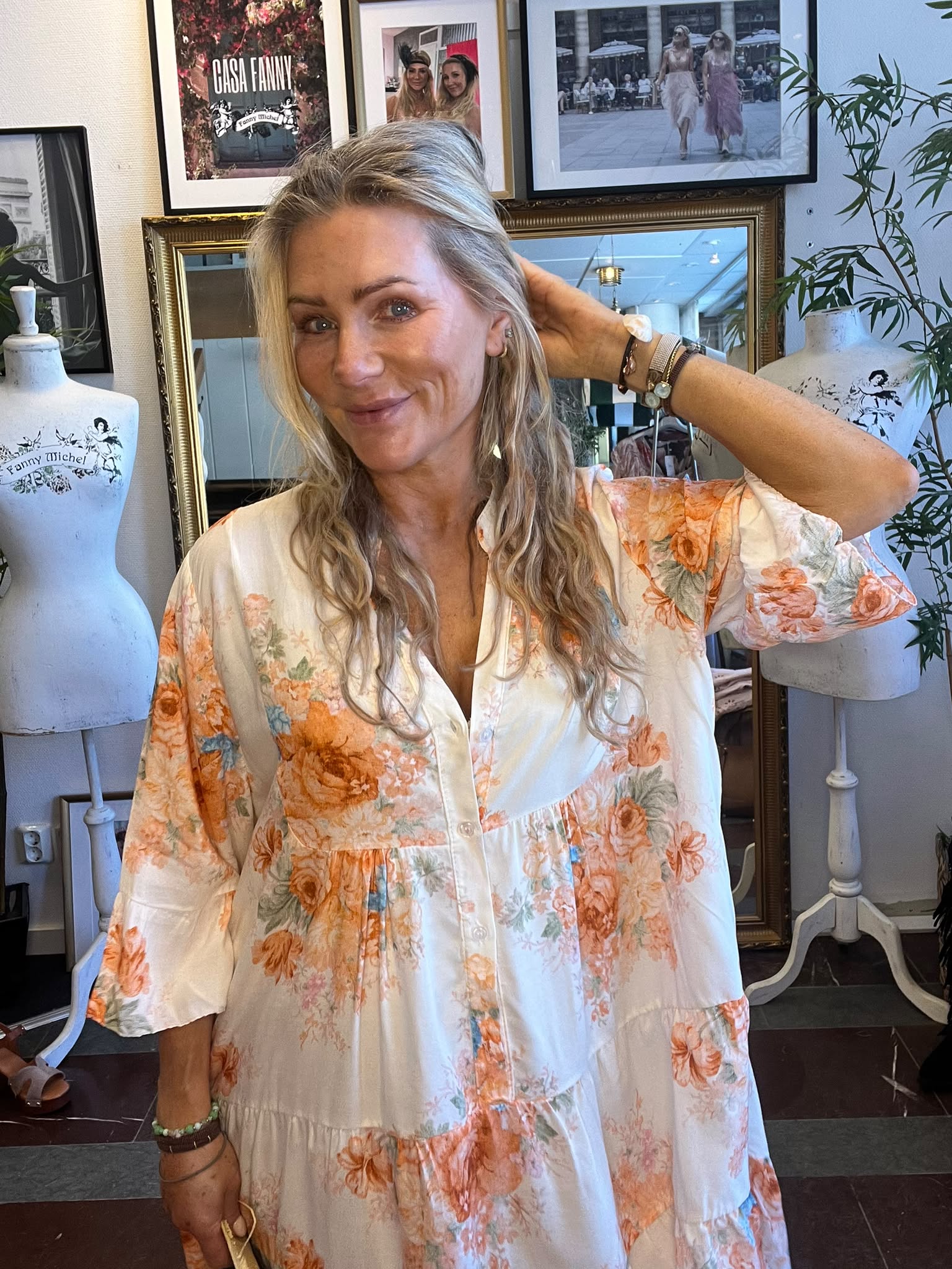 Leende kvinna i vit klänning med orange blommönster håller handen bakom huvudet och poserar inomhus. Bakom henne syns provdockor och inramade foton, samt texten “CASA FANNY” och “Fanny Michel”.