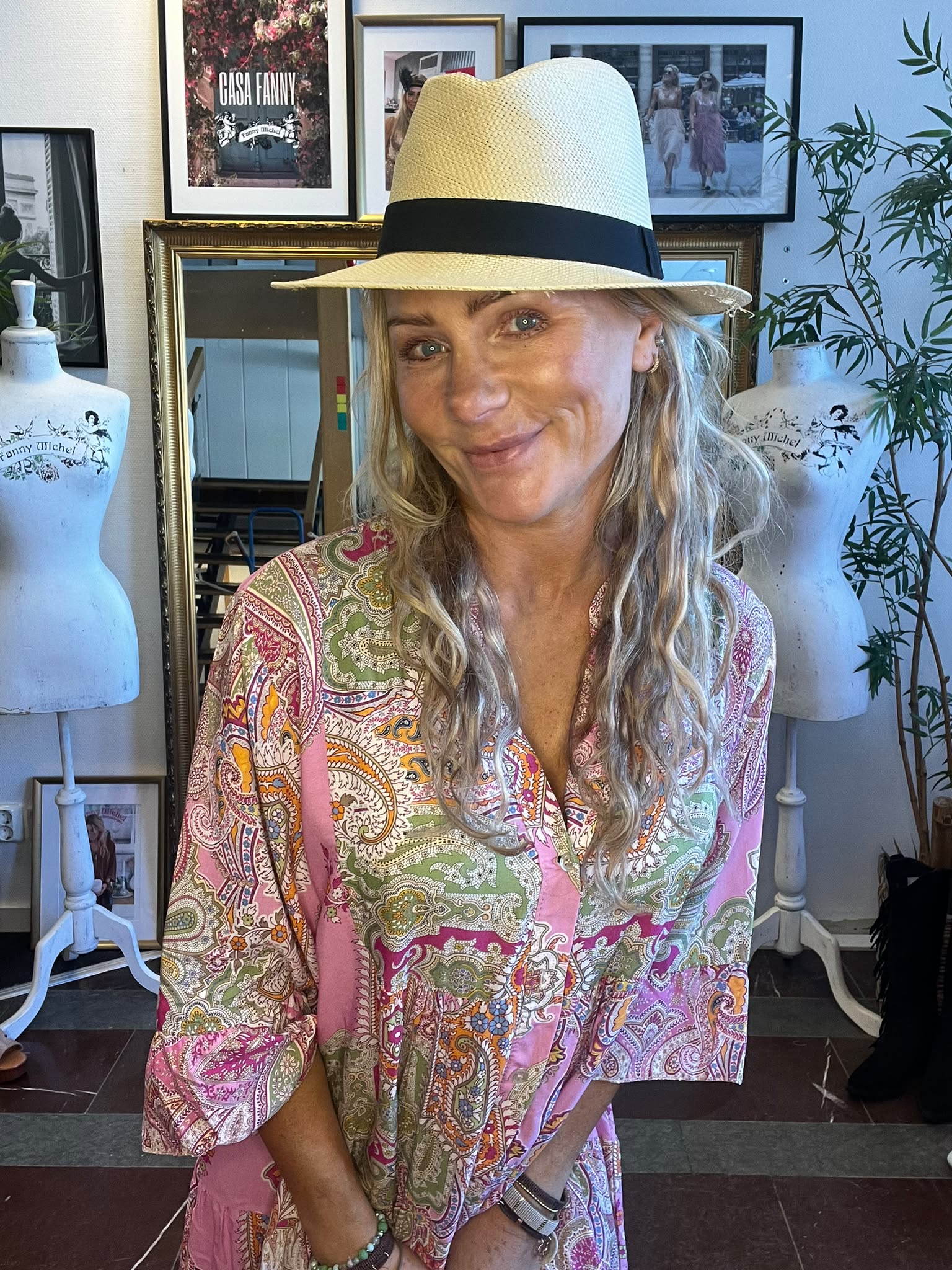 Leende kvinna i halmhatt och färgglad paisleymönstrad blus poserar framför en vägg med inramade foton. I bakgrunden syns provdockor med trycket “Fanny Michel” samt en tavla med texten “CASA FANNY”.