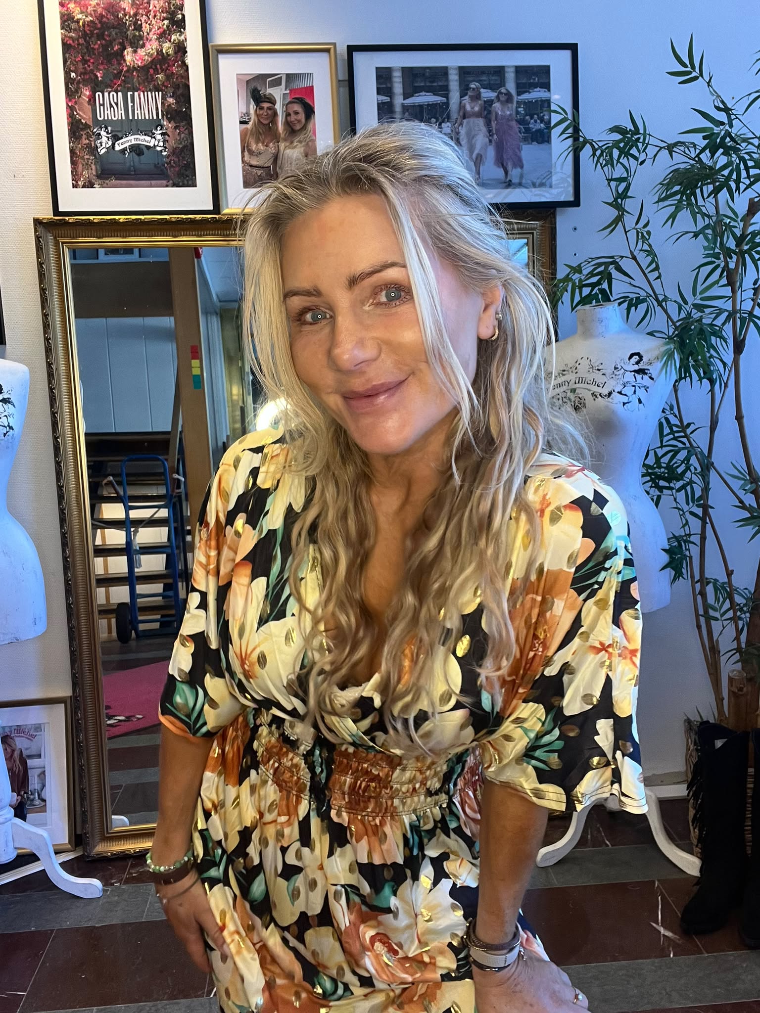 Blond kvinna i blommig klänning lutar sig fram och ler mot kameran i ett rum med guldspegel, provdockor och en stor växt. På väggen syns inramade foton, varav ett med texten “CASA FANNY”.