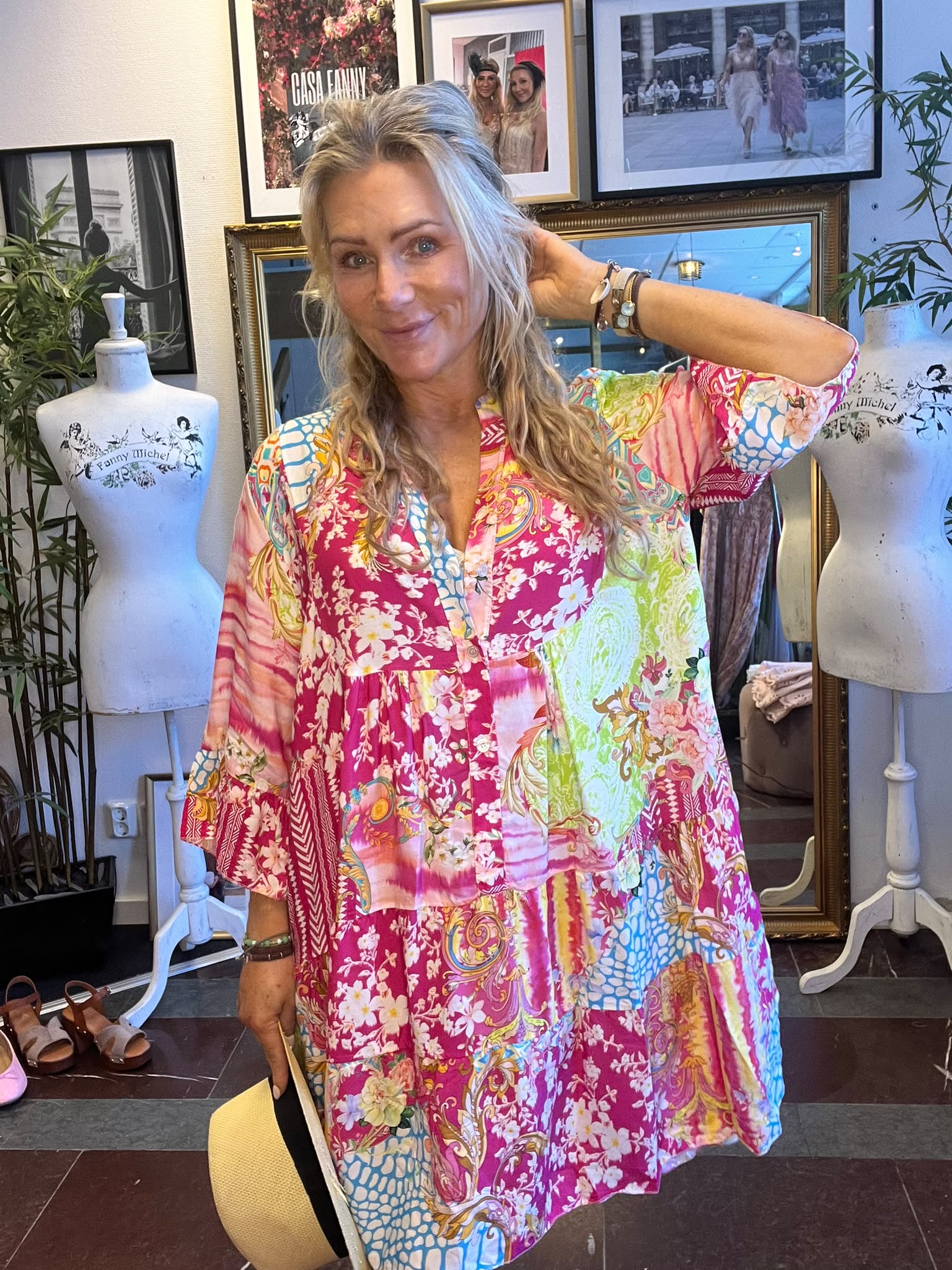 Blond kvinna i färgstark, mönstrad klänning poserar och håller handen bakom huvudet i en butik. Runt henne står provdockor med texten “Fanny Michel”, och på väggen hänger inramade foton, bland annat “CASA FANNY”.