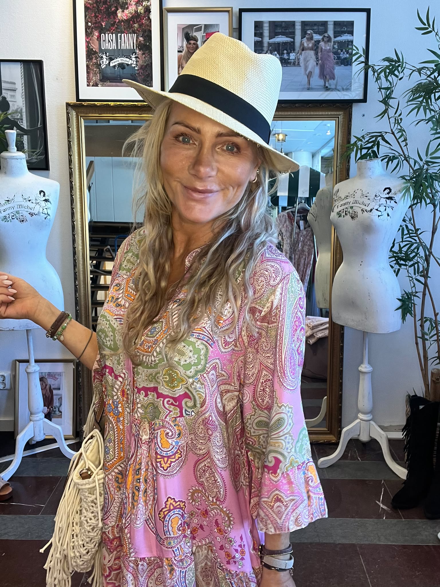 Leende kvinna i stråhatt och rosa mönstrad tunika poserar och håller i en skyltdocka i en butik/ateljé. Bakom syns spegel, väggfoton och skyltdockor med texten “Fanny Michel” samt en affisch med “CASA FANNY”.