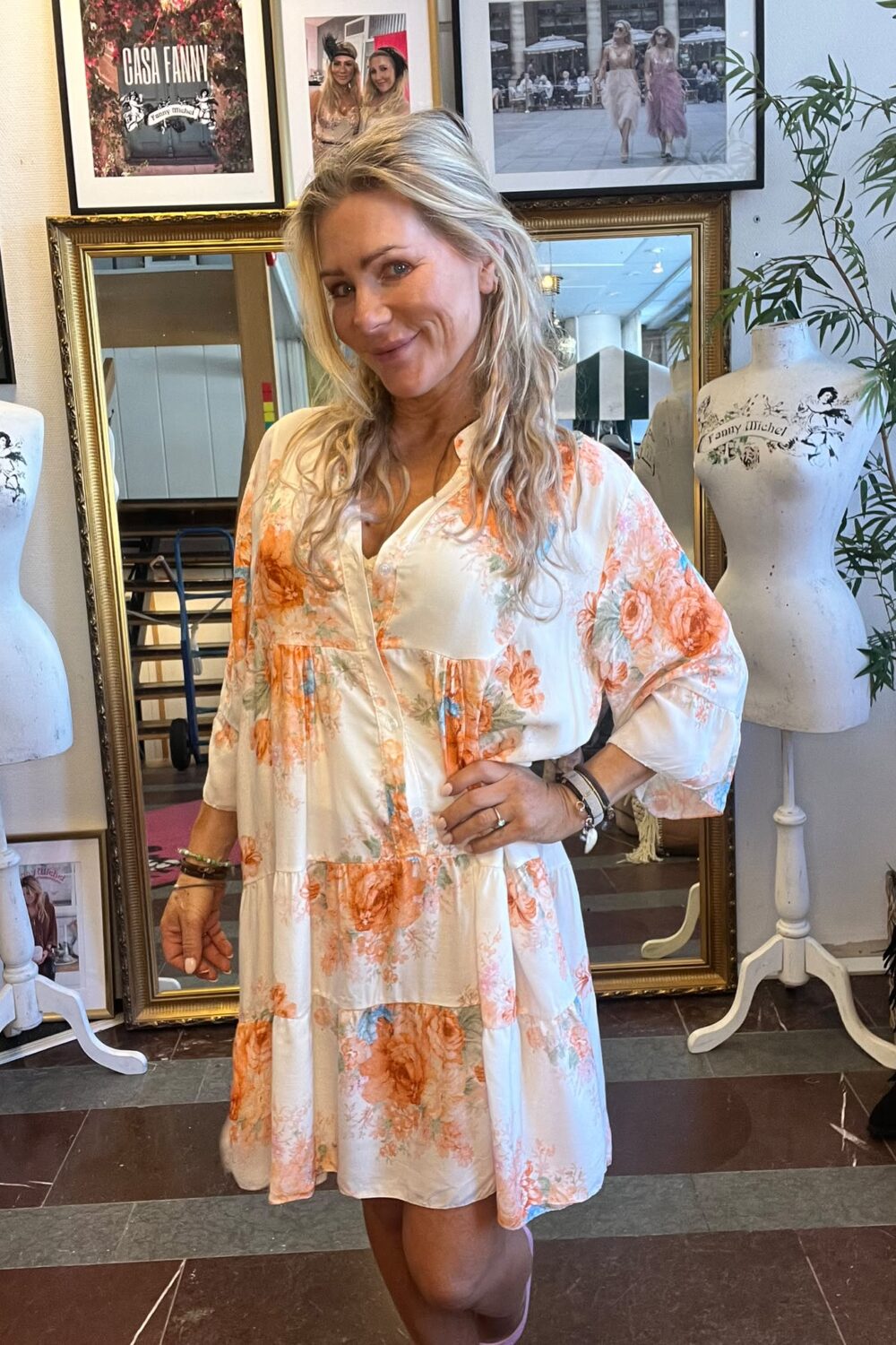 Blond kvinna poserar leende med handen på höften i en blommig vit klänning, framför en stor guldramsspegel. Runt henne står två provdockor med texten “Fanny Michel”; på väggen syns foton och skylten “CASA FANNY”.