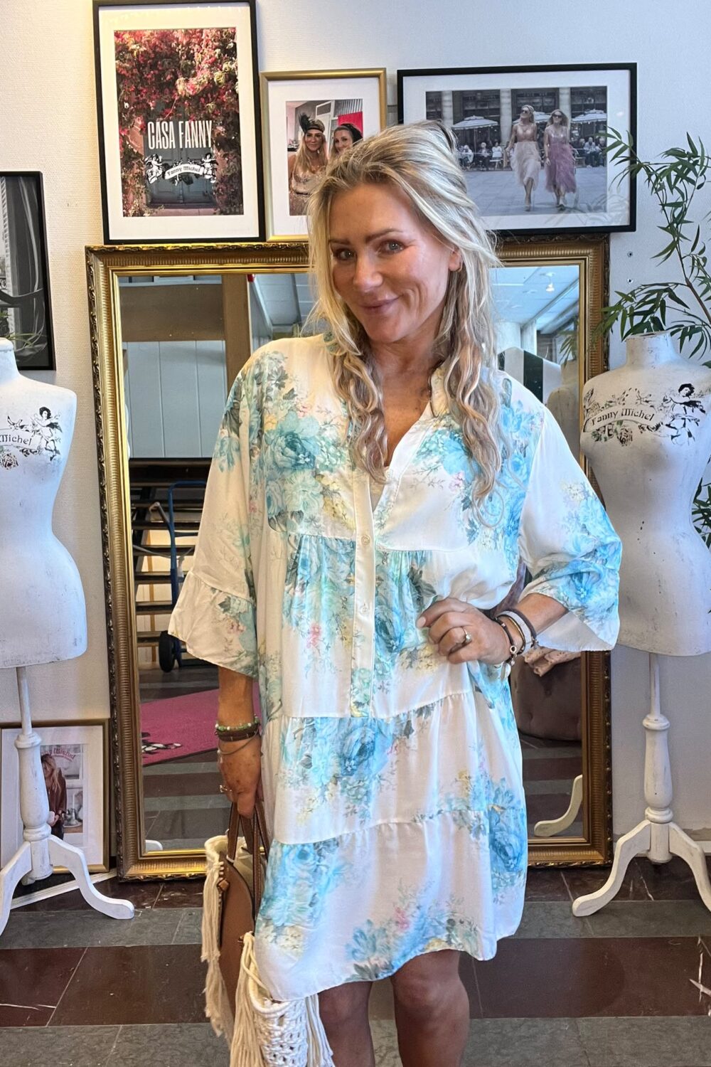Leende kvinna i ljusblå blommig klänning står och poserar med handväska framför en stor guldramsspegel i en butik. Runt henne syns provdockor med texten ”Fanny Michel” och inramade foton, inklusive ”CASA FANNY”.