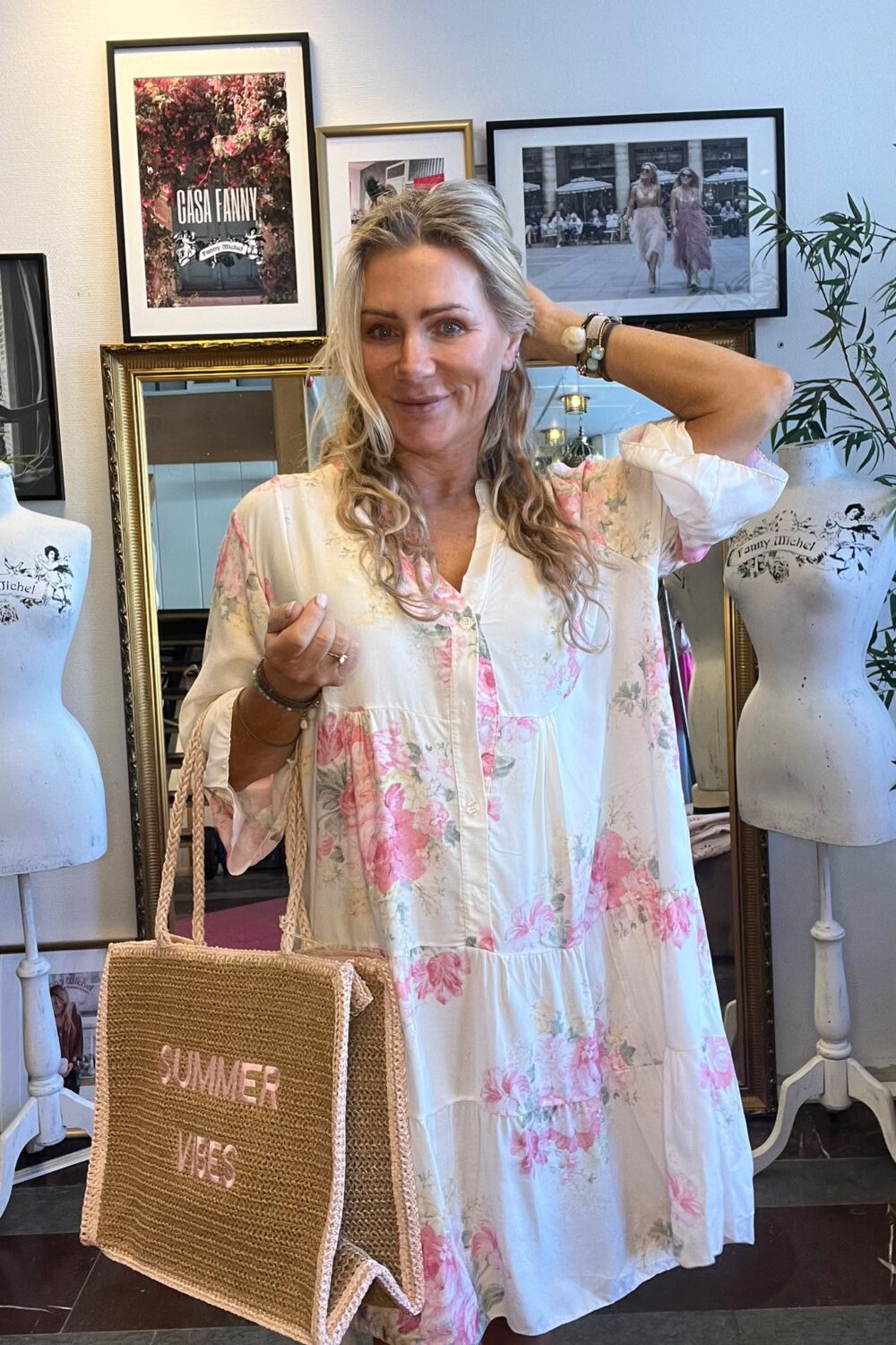 Leende kvinna i vit klänning med rosa blommor poserar inomhus, med ena handen bakom huvudet och en halmväska med texten “SUMMER VIBES” i andra. Bakom syns provdockor och inramade bilder, bland annat “CASA FANNY”.