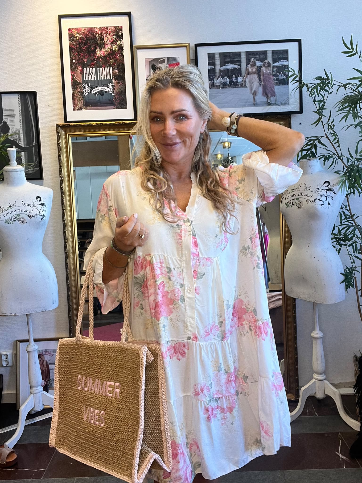 Leende kvinna i vit klänning med rosa blommor poserar inomhus, med ena handen bakom huvudet och en halmväska med texten “SUMMER VIBES” i andra. Bakom syns provdockor och inramade bilder, bland annat “CASA FANNY”.