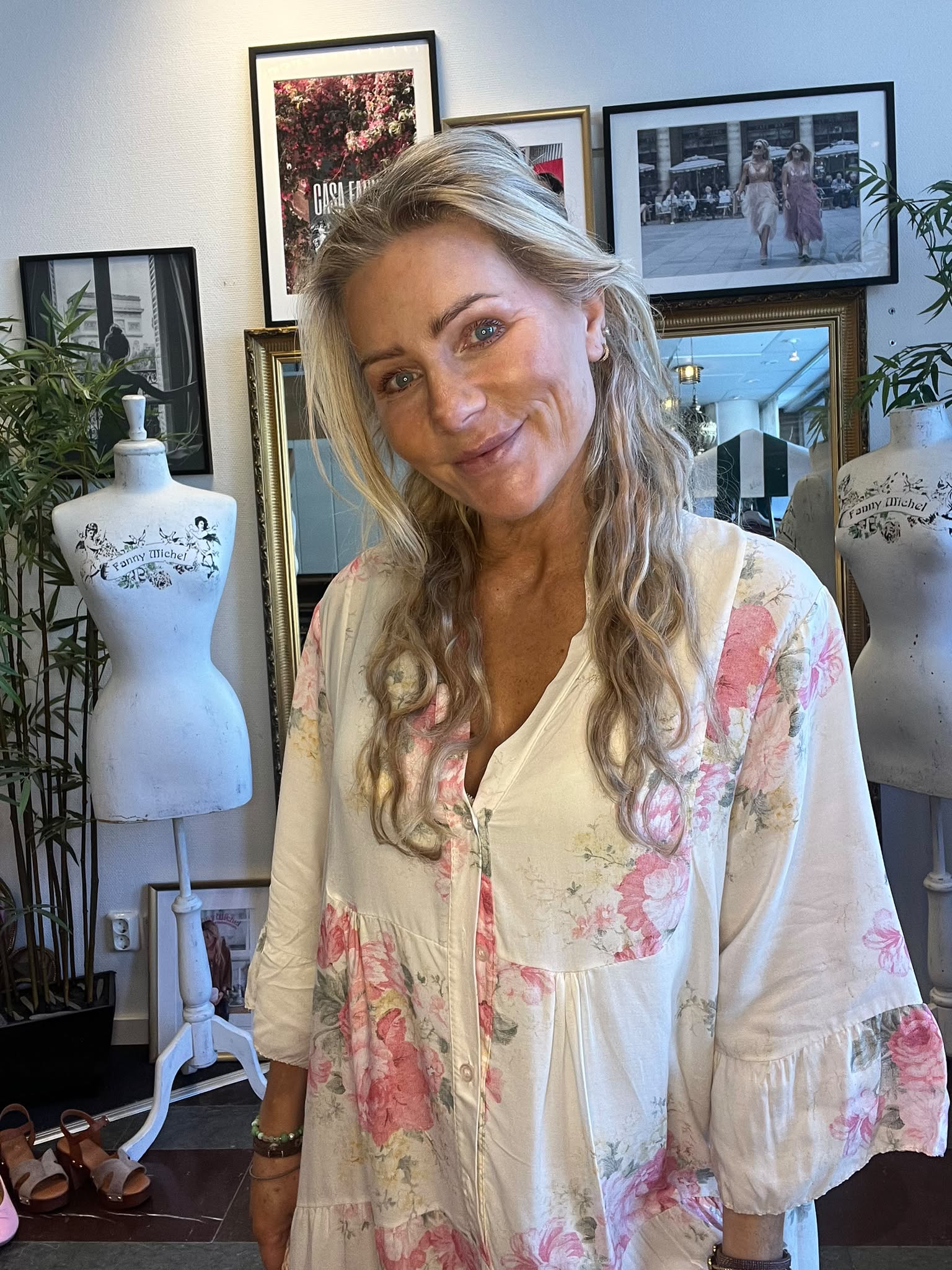 Leende kvinna med långt blont hår lutar huvudet mot kameran och bär en vit klänning med rosa blommor. Hon står i en ateljélik miljö med inramade foton och provdockor märkta “Fanny Michel”.