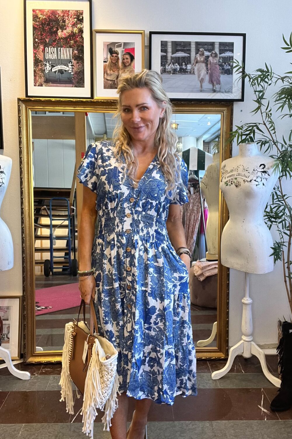 Leende kvinna i blåvit blommig klänning håller en brun väska med vita fransar i en klädbutik. Bakom henne syns en stor guldramad spegel, provdockor och inramade foton; text: “CASA FANNY”.