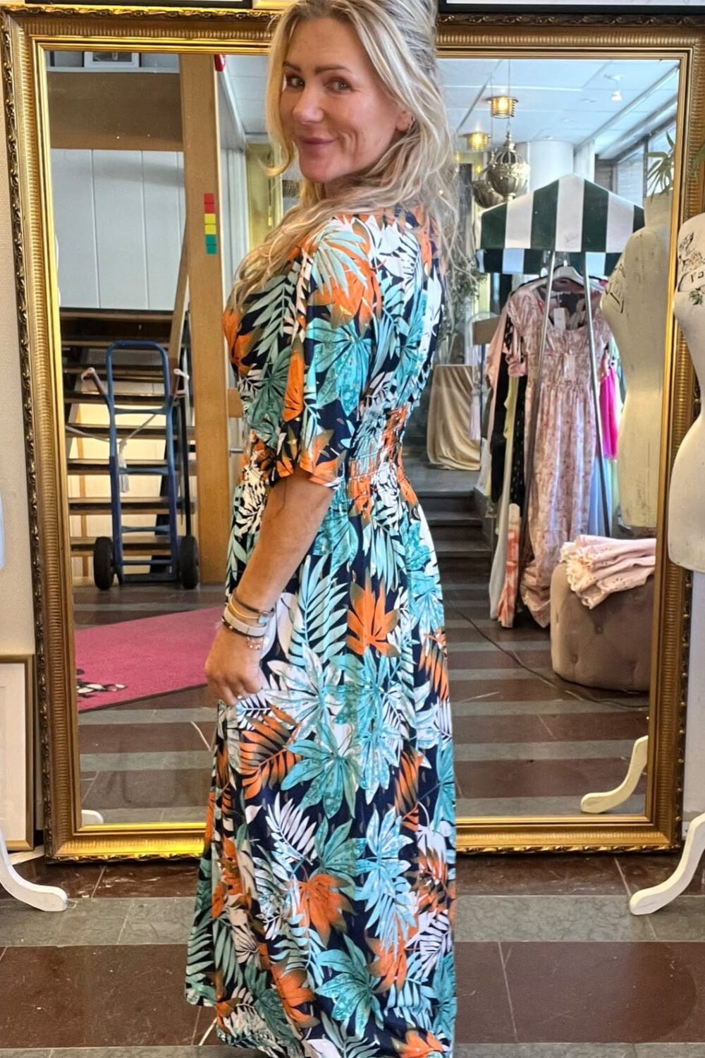 Blond kvinna står framför en stor guldramsspegel och vänder sig leende mot kameran. Hon bär en lång blommig maxiklänning i grönt och orange samt vita sneakers. I bakgrunden syns klädhängare och provrumsbutik.