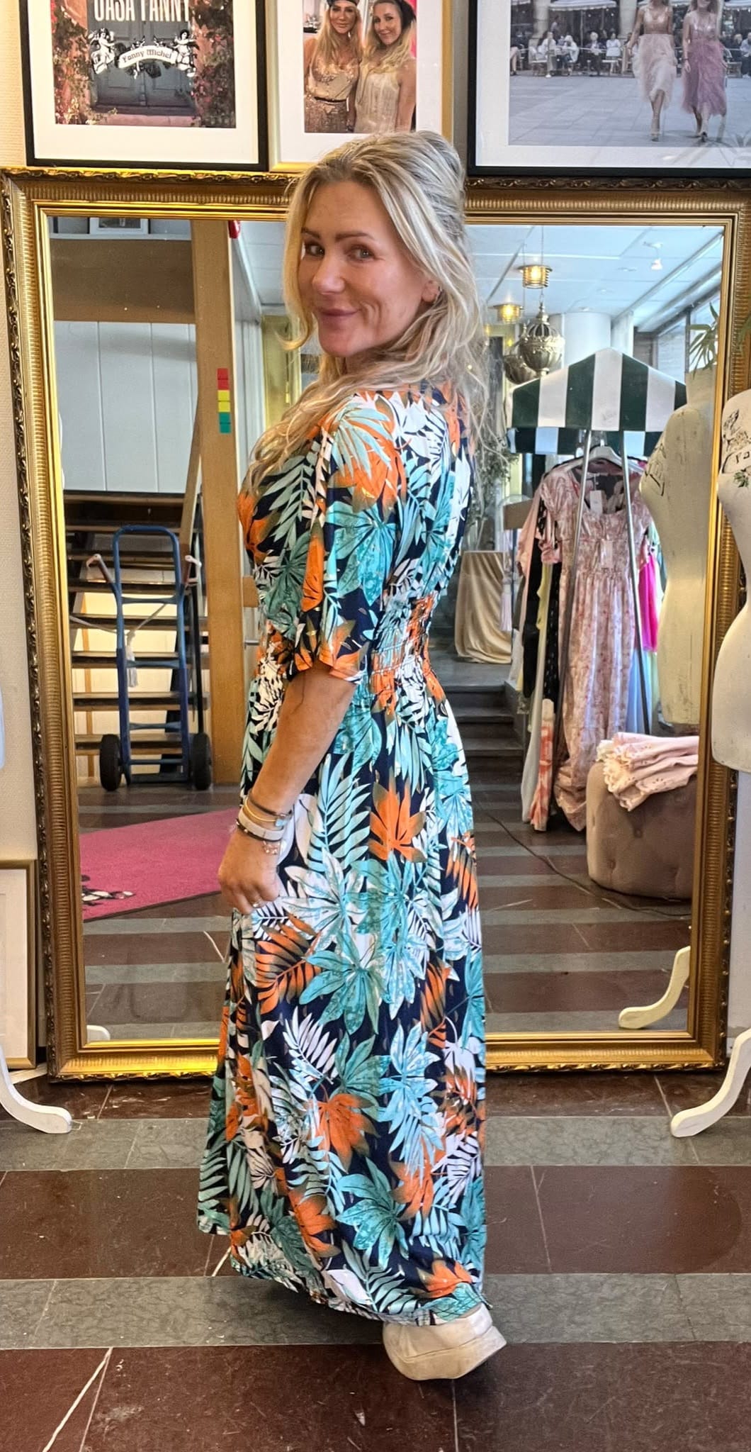 Blond kvinna står framför en stor guldramsspegel och vänder sig leende mot kameran. Hon bär en lång blommig maxiklänning i grönt och orange samt vita sneakers. I bakgrunden syns klädhängare och provrumsbutik.