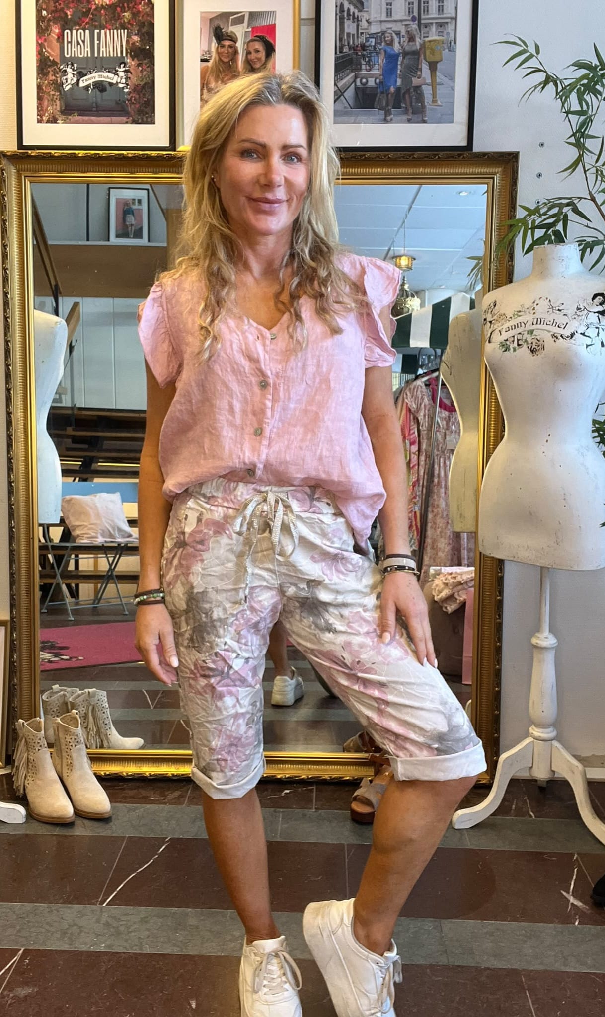 Leende kvinna poserar i en klädbutik framför en stor guldramad spegel, klädd i rosa blus, blommiga capribyxor och vita sneakers. Bredvid står skyltdockor; på väggen syns texten “CASA FANNY” och “Fanny Michel”.