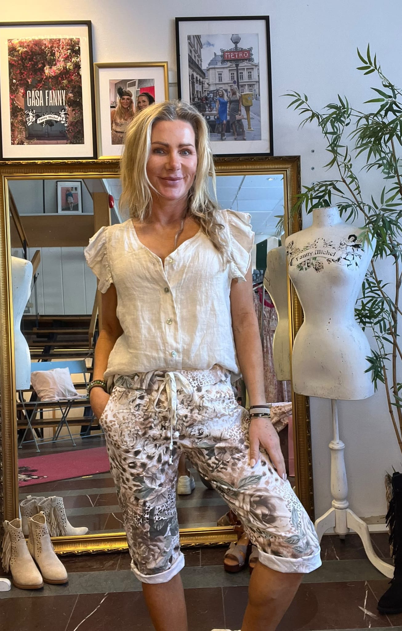 Kvinna med blont hår poserar och ler i en butik/ateljé, iklädd vit blus och mönstrade byxor. Bakom syns spegel, skyltdocka och inramade foton med texten “CASA FANNY” och “METRO”.