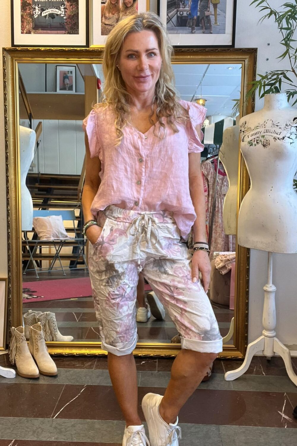 Leende kvinna står med händerna i fickorna framför en stor guldramsspegel i en klädbutik, klädd i rosa blus och blommiga shorts med vita sneakers. I bakgrunden syns skyltdockor och inramade foton med texten ”CASA FANNY” och ”Fanny Michel”.