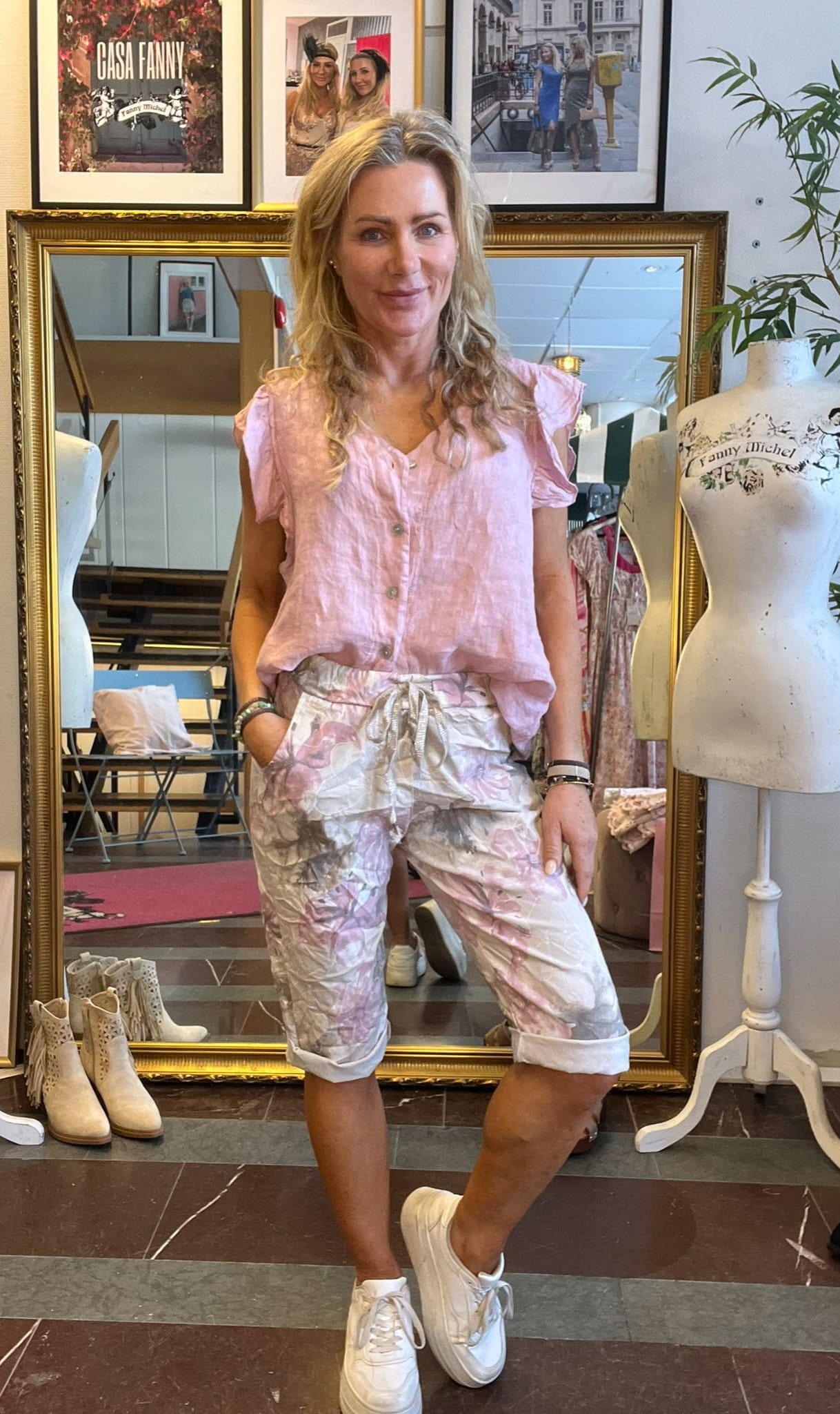 Leende kvinna står med händerna i fickorna framför en stor guldramsspegel i en klädbutik, klädd i rosa blus och blommiga shorts med vita sneakers. I bakgrunden syns skyltdockor och inramade foton med texten ”CASA FANNY” och ”Fanny Michel”.
