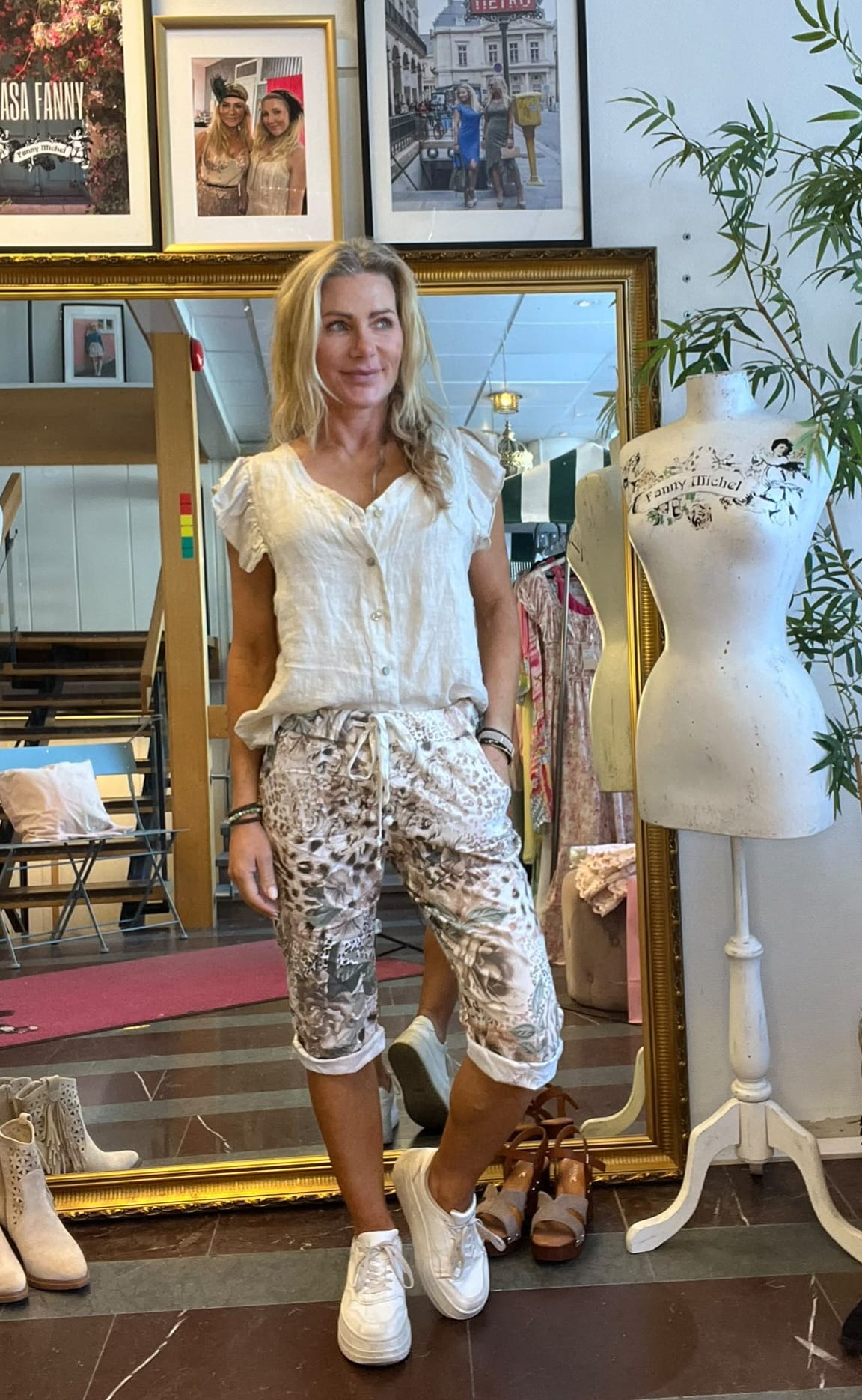 Blond kvinna står avslappnat med händerna i fickorna framför en stor guldramsspegel i en klädbutik, klädd i vit blus, mönstrade capribyxor och vita sneakers. Bredvid syns provdocka med texten “Fanny Michel” och skor på golvet.