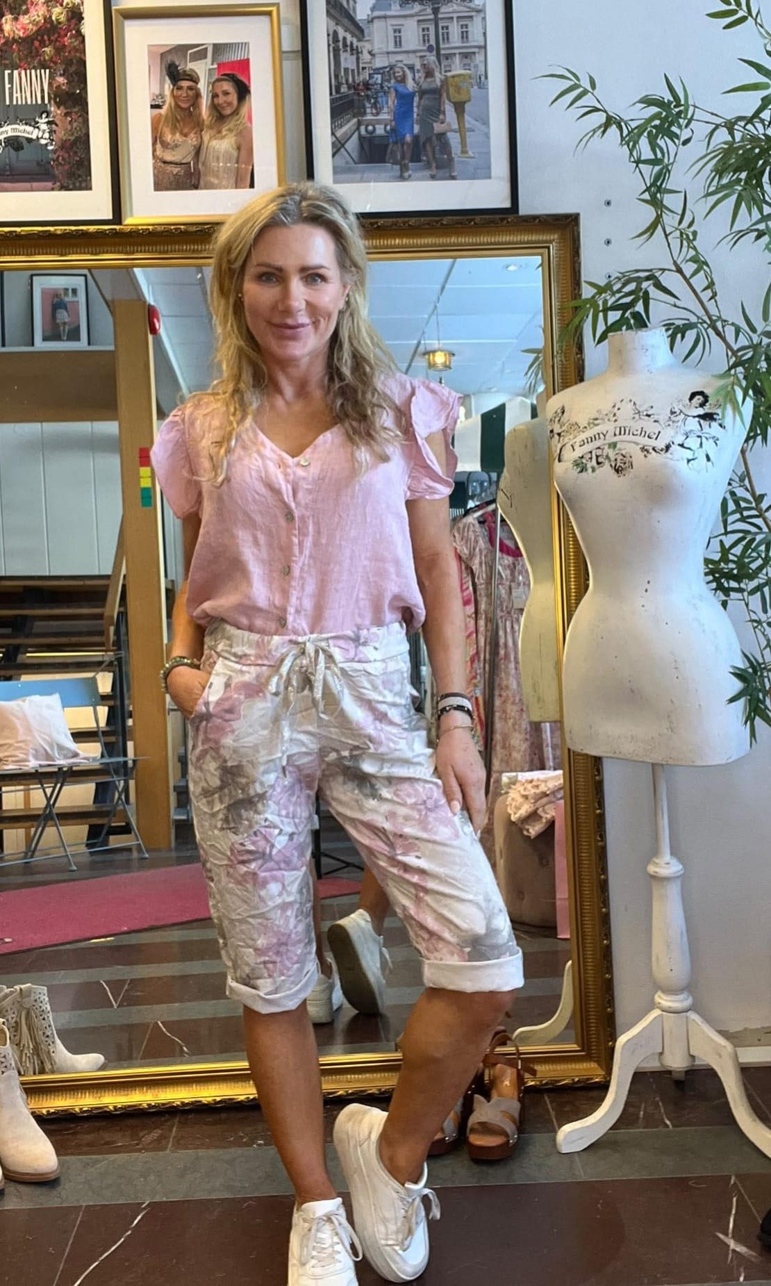 Leende kvinna poserar framför en stor guldramsspegel i en klädbutik, klädd i ljusrosa blus och ljusa mönstrade capribyxor med vita sneakers. Bredvid står provdocka med texten ”Fanny Michel” och inramade foton ovanför.