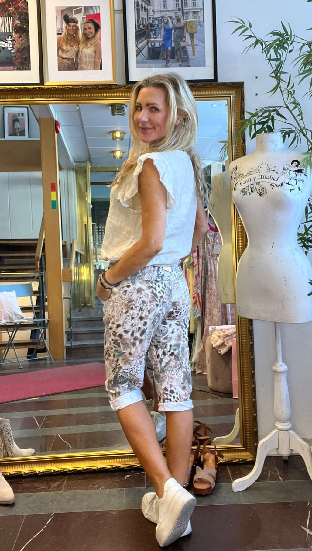 Blond kvinna står vänd i profil och ler mot kameran, med händerna i fickorna på mönstrade capribyxor och vit blus. Hon befinner sig i en butik med stor guldramad spegel, foton på väggen och provdocka med texten ”Lanny Michel”.
