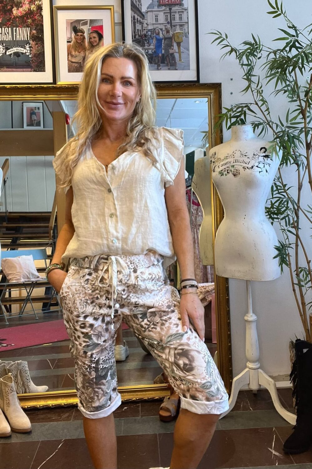 Kvinna står och ler i en klädbutik, med handen i fickan, klädd i vit blus och mönstrade capribyxor. Bakom henne syns spegel, inramade foton med texten “CASA FANNY” och en provdocka märkt “Fanny Michel”.