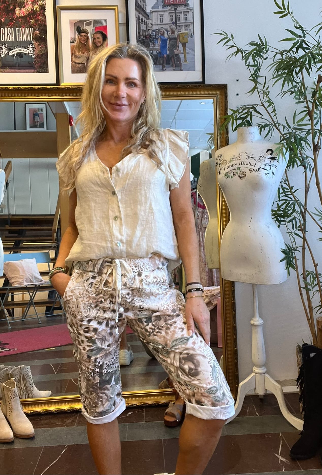 Leende kvinna poserar med handen i fickan i en klädbutik, klädd i vit blus och mönstrade capribyxor. Bakom henne syns stora inramade foton med texten “CASA FANNY” samt en provdocka märkt “Fanny Michel”.