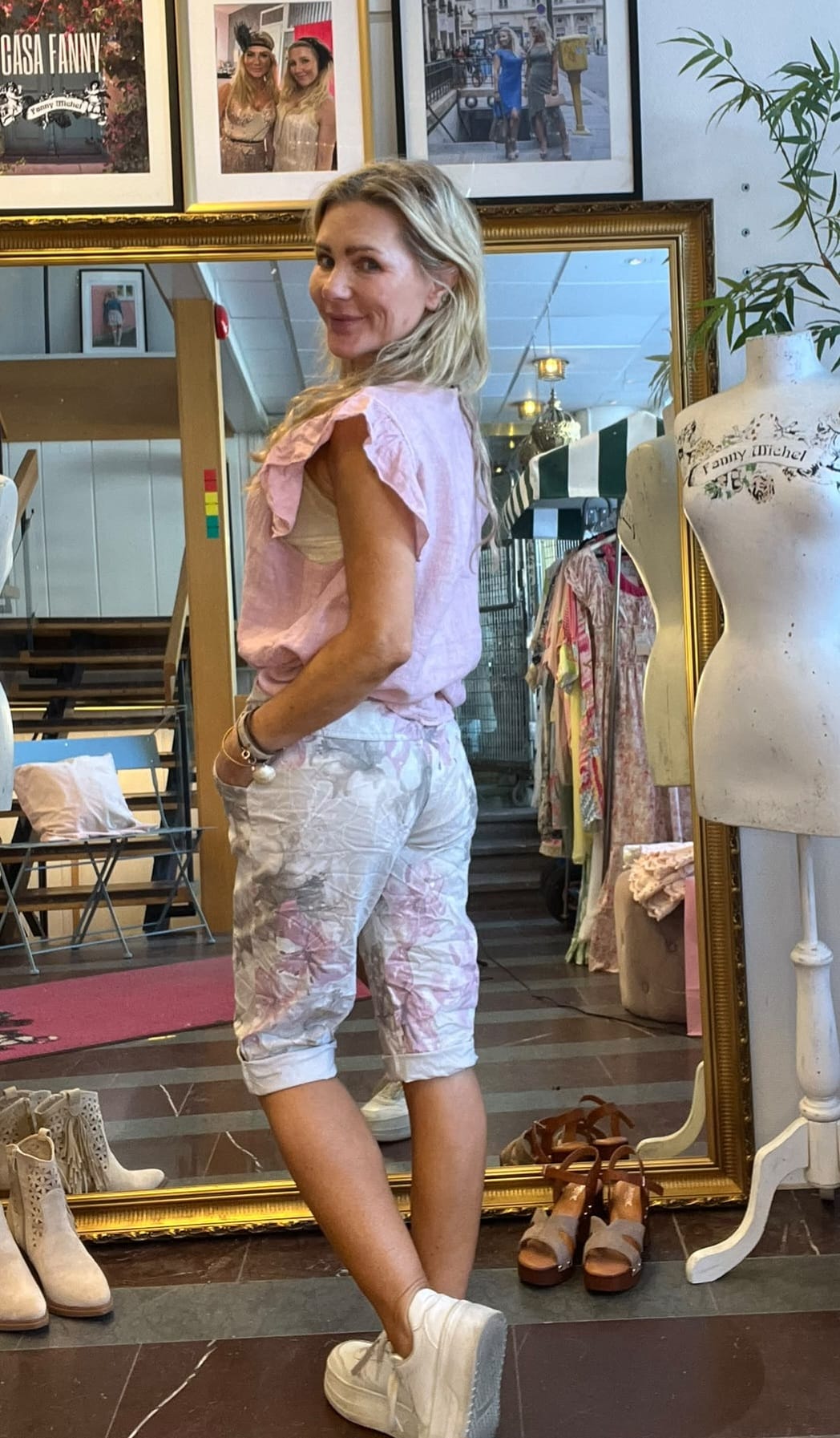 Kvinna står i en klädbutik och poserar framför en stor guldramsspegel, vänd halvvägs mot kameran med ett leende. Hon bär rosa topp och mönstrade shorts. Intill syns provdocka med texten “Fanny Michel” och skor på golvet.