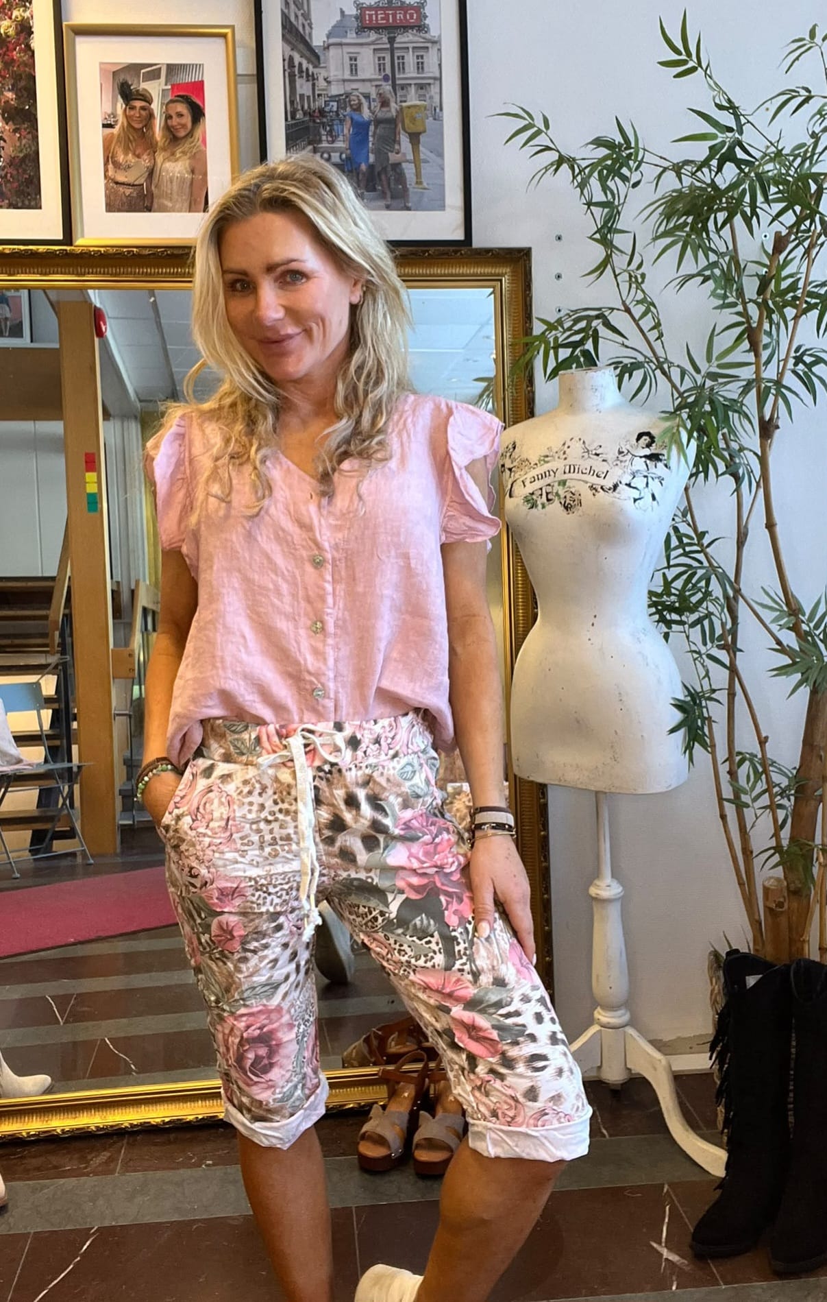 Blond kvinna poserar och ler med händerna i fickorna, iklädd ljusrosa blus och mönstrade capribyxor. Hon står i en butik framför speglar och inramade foton; bredvid syns provdocka med texten “Fanny Michel” och en grön växt.