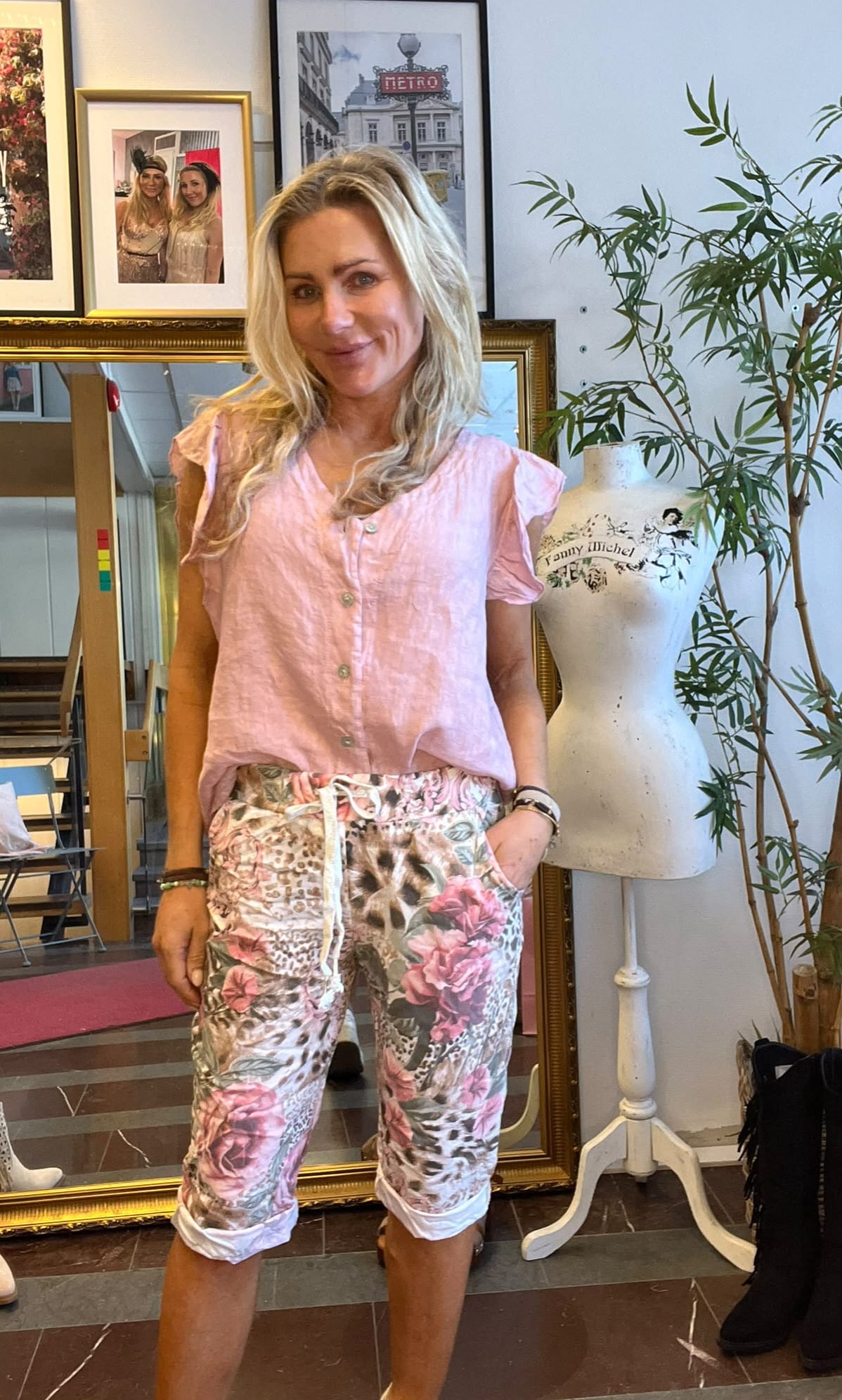 Leende kvinna med långt blont hår poserar inomhus med händerna i fickorna, klädd i ljusrosa blus och blommiga capribyxor. Bakom syns inramade foton och speglar, samt en provdocka med texten “Fanny Michel”.