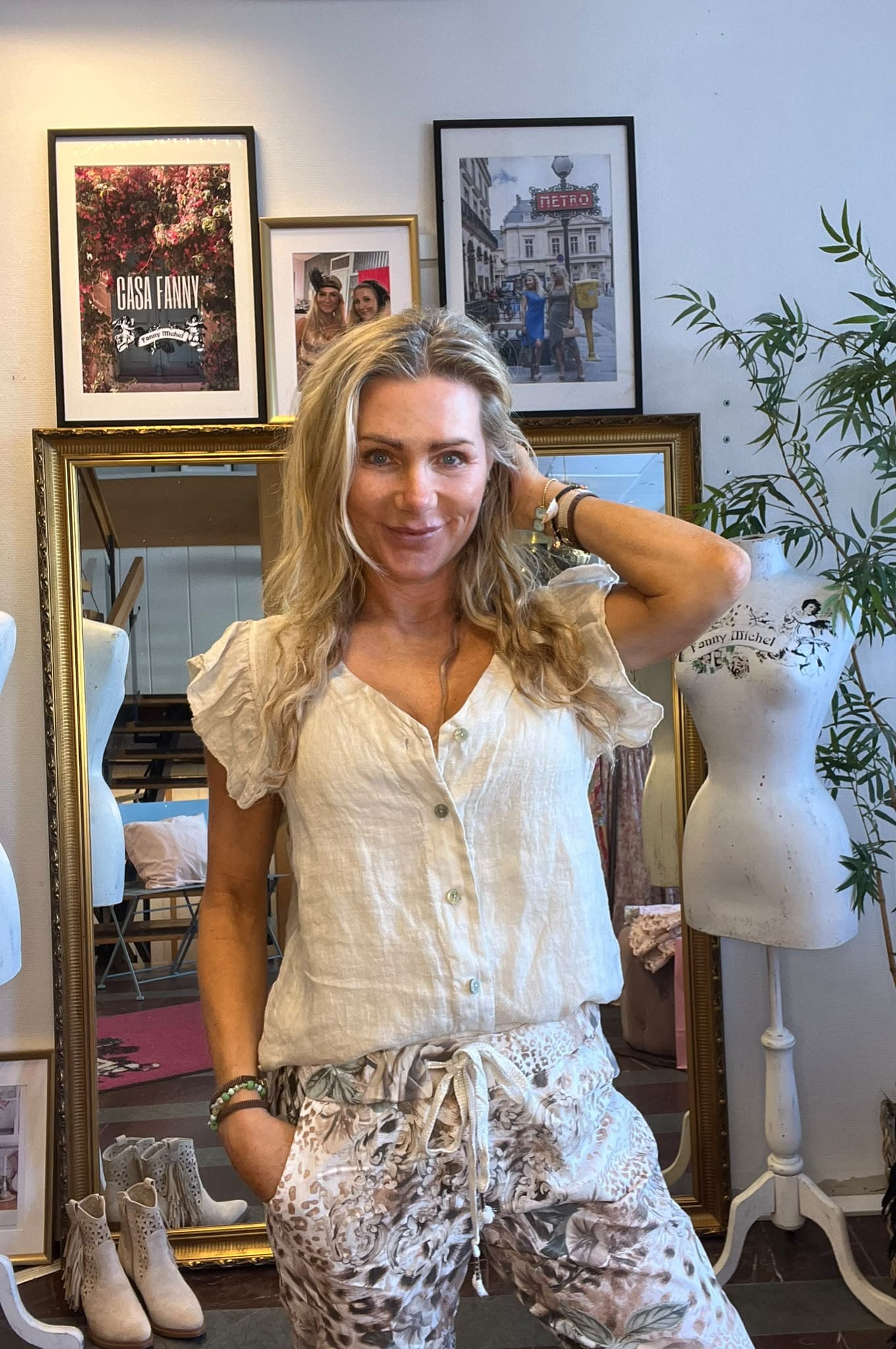 Leende kvinna i ljus blus poserar med ena handen bakom huvudet i en butik/ateljé. Bakom henne syns en spegel, provdockor och inramade bilder med texten “CASA FANNY” och “METRO”, samt en grön växt.