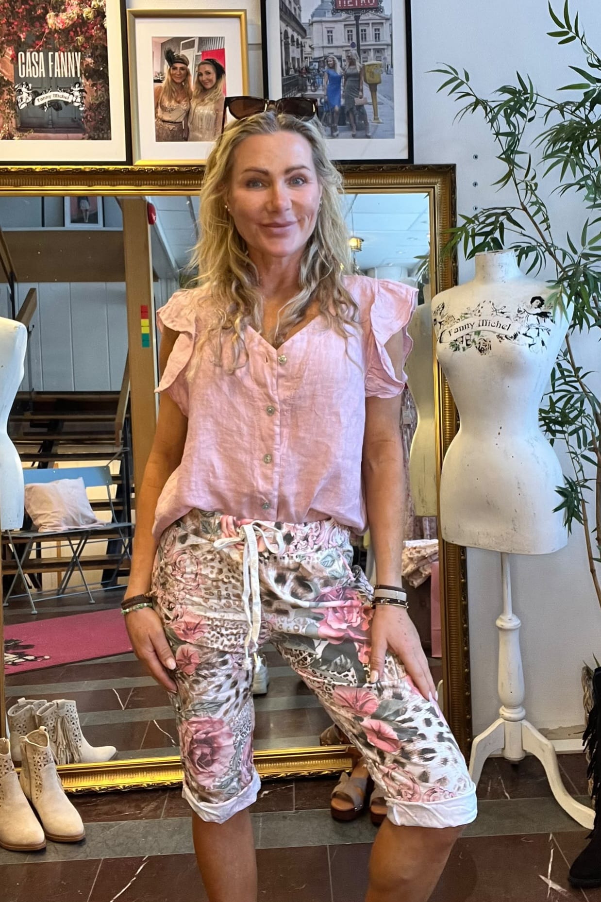 Kvinna poserar leende framför en stor guldramsspegel i en butik, iklädd ljusrosa blus och blommiga capribyxor. Runt henne syns skor, en skyltdocka med texten ”Fanny Michel” och inramade foton, varav ett säger ”CASA FANNY”.