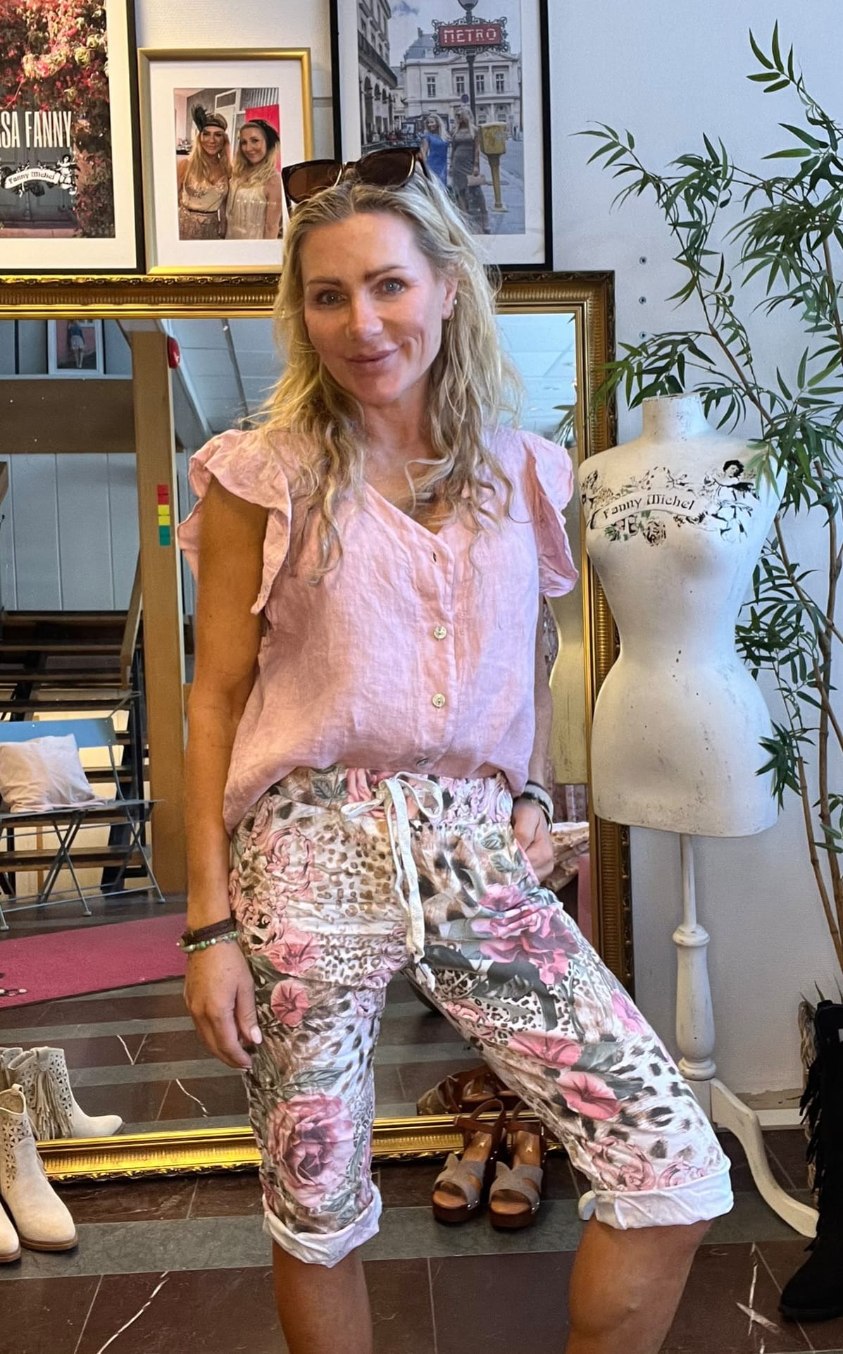 Blond kvinna poserar och ler i en butik/ateljé, klädd i ljusrosa blus och blommiga capribyxor med solglasögon på huvudet. Bredvid står en provdocka med texten “Fanny Michel”; ramar och skor syns i bakgrunden.