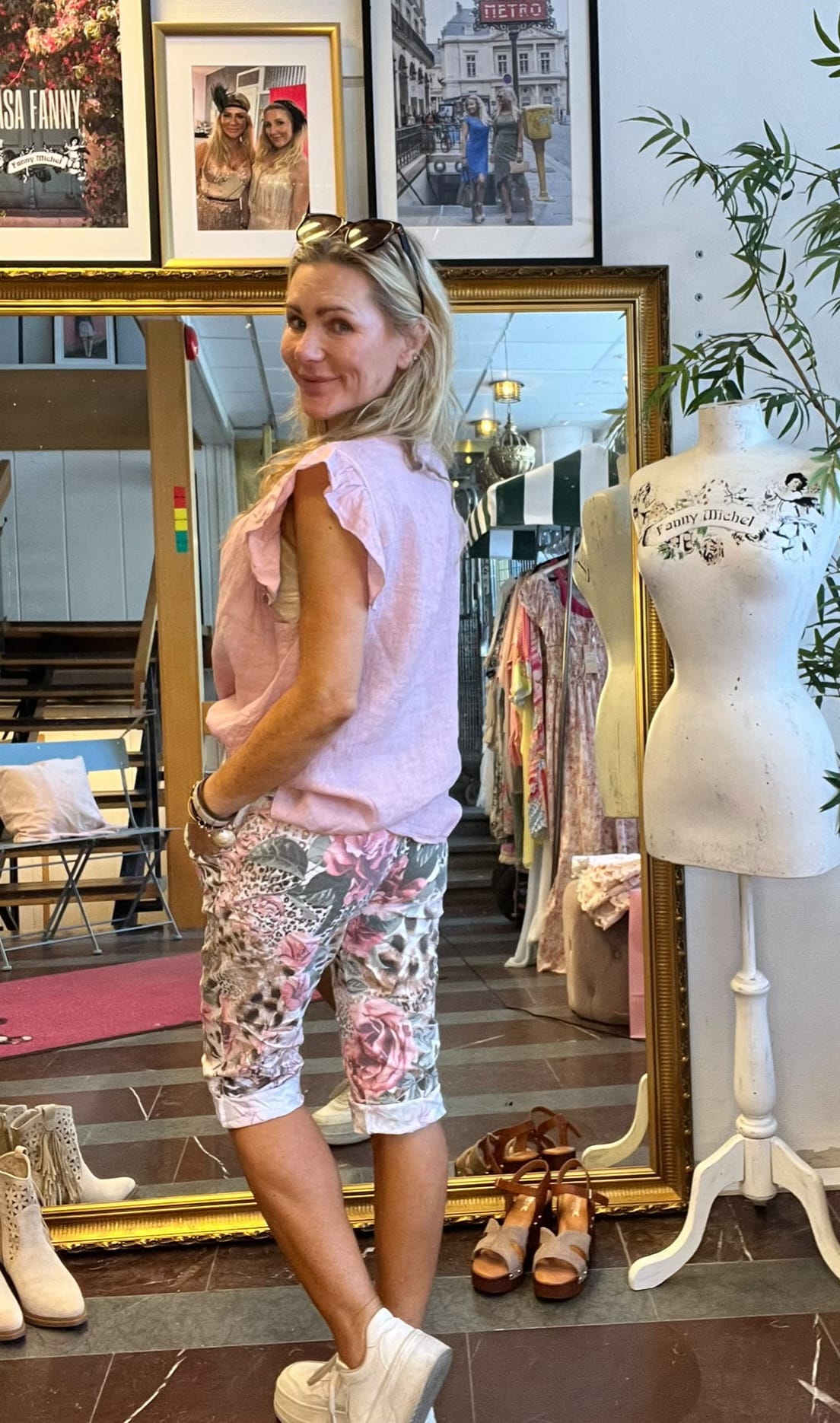 Kvinna i rosa blus och blommiga byxor står och ler mot kameran i en klädbutik. Bakom henne speglar en stor guldram spegel lokalen; intill syns provdocka med texten ”Fanny Michel” samt kläder och skor.