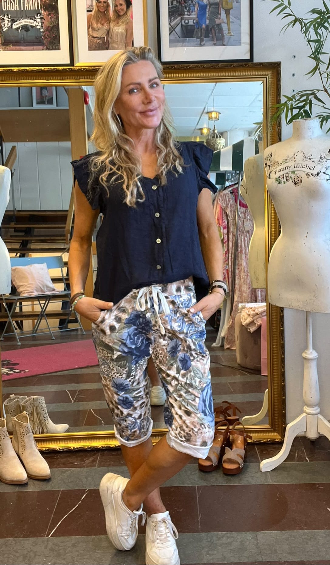 Blond kvinna poserar med händerna i fickorna framför en stor guldramad spegel i en klädbutik, klädd i mörk blus, blommiga capribyxor och vita sneakers; intill står en provdocka med texten “Fanny Michel”.