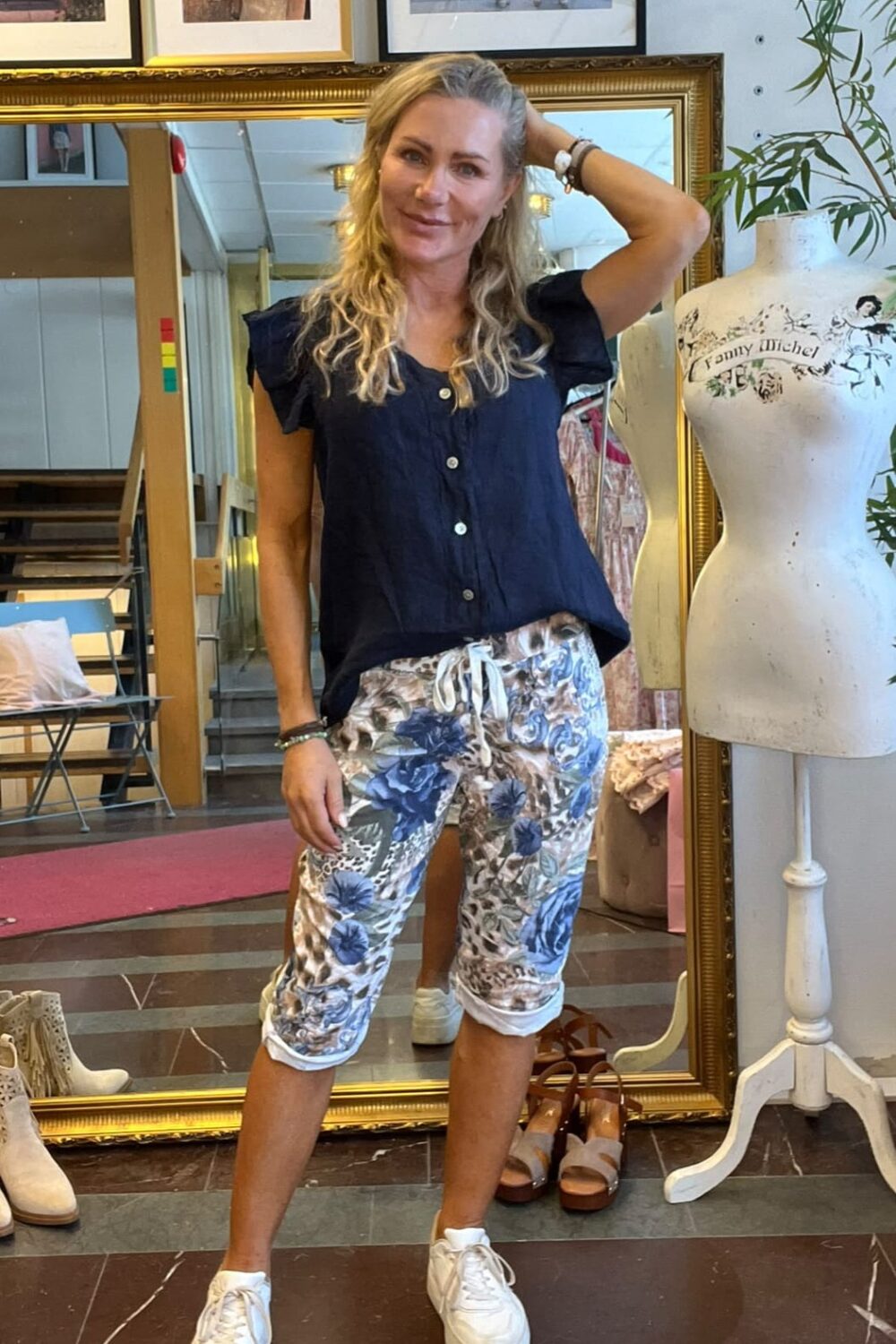 Kvinna med långt blont hår poserar med ena handen bakom huvudet och ler, iklädd mörkblå topp, mönstrade capribyxor och vita sneakers. Hon står i en butik framför en guldramsspegel bredvid en provdocka märkt “Fanny Michel”.