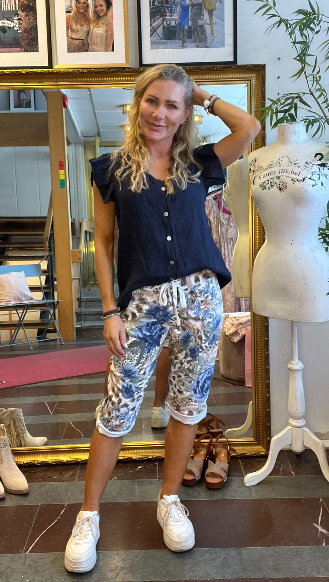 Kvinna med långt blont hår poserar med ena handen bakom huvudet och ler, iklädd mörkblå topp, mönstrade capribyxor och vita sneakers. Hon står i en butik framför en guldramsspegel bredvid en provdocka märkt “Fanny Michel”.
