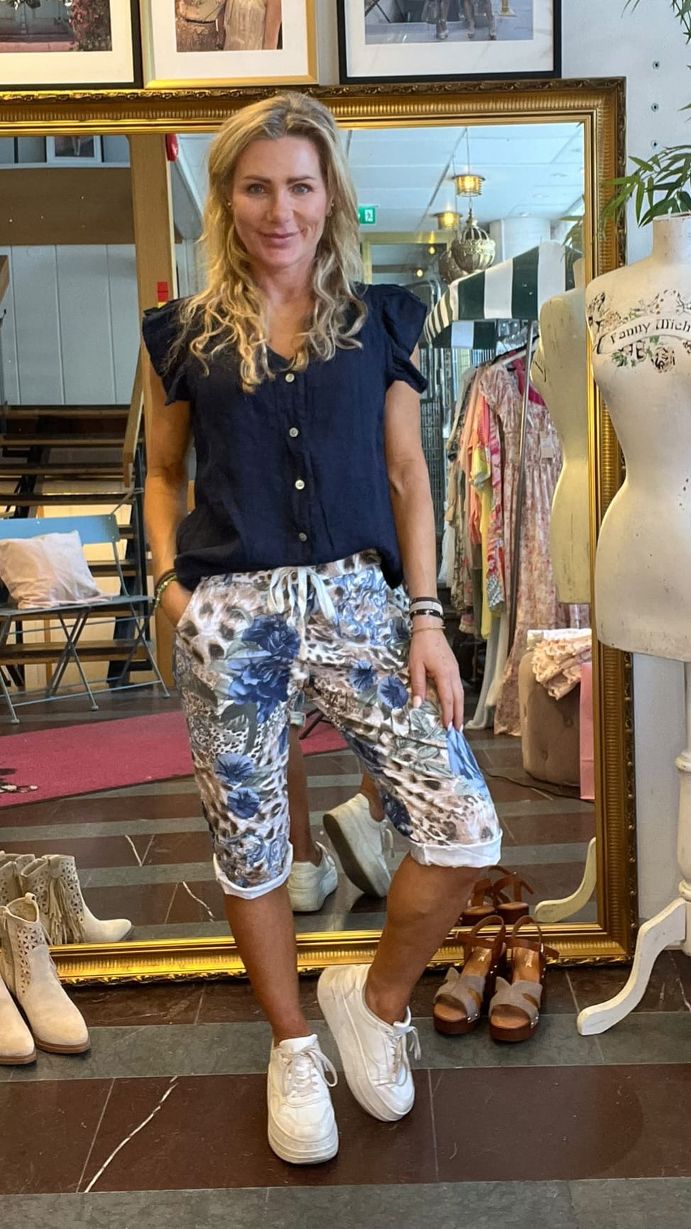 Kvinna med långt blont hår poserar framför en stor guldramsspegel i en klädbutik, med ena benet böjt. Hon bär mörkblå topp, blommiga capribyxor och vita sneakers; klädställningar, skor och en provdocka syns.
