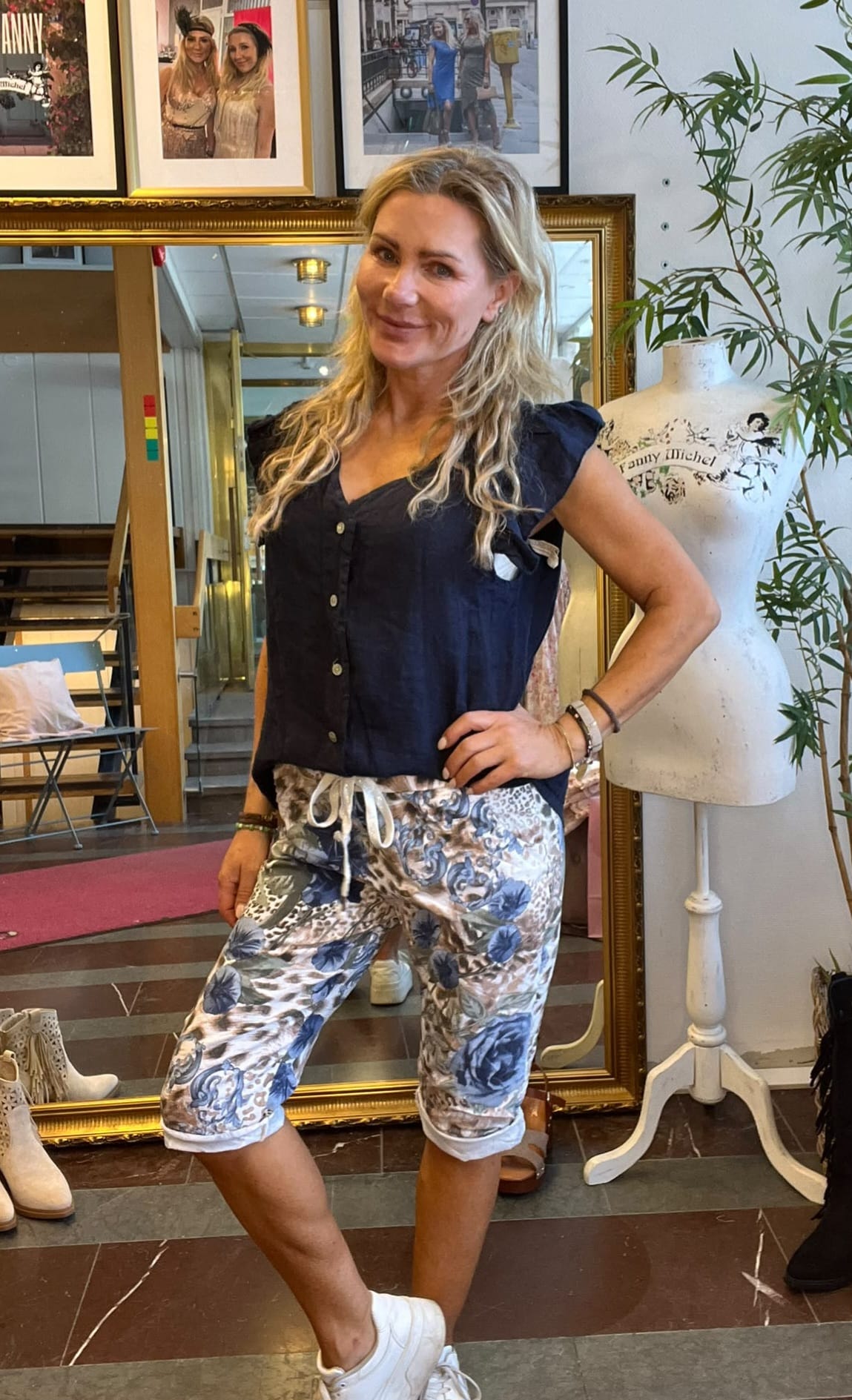 Kvinna står och poserar med handen på höften i en klädbutik, iklädd mörk blus och blommiga capribyxor med snörning. Bakom syns guldramsspegel, inramade foton samt provdocka med texten “Fanny Michel”.