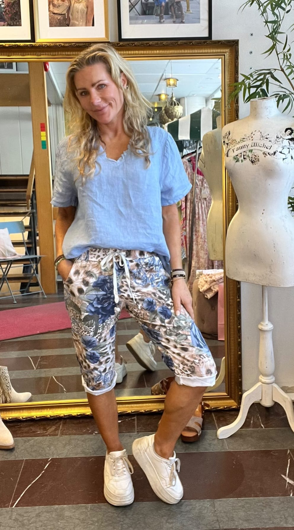 Leende kvinna poserar framför en stor guldramsspegel i en klädbutik, med handen i fickan. Hon bär ljusblå topp, mönstrade capribyxor och vita sneakers. Bredvid står en provdocka med texten ”Fanny Michel”.