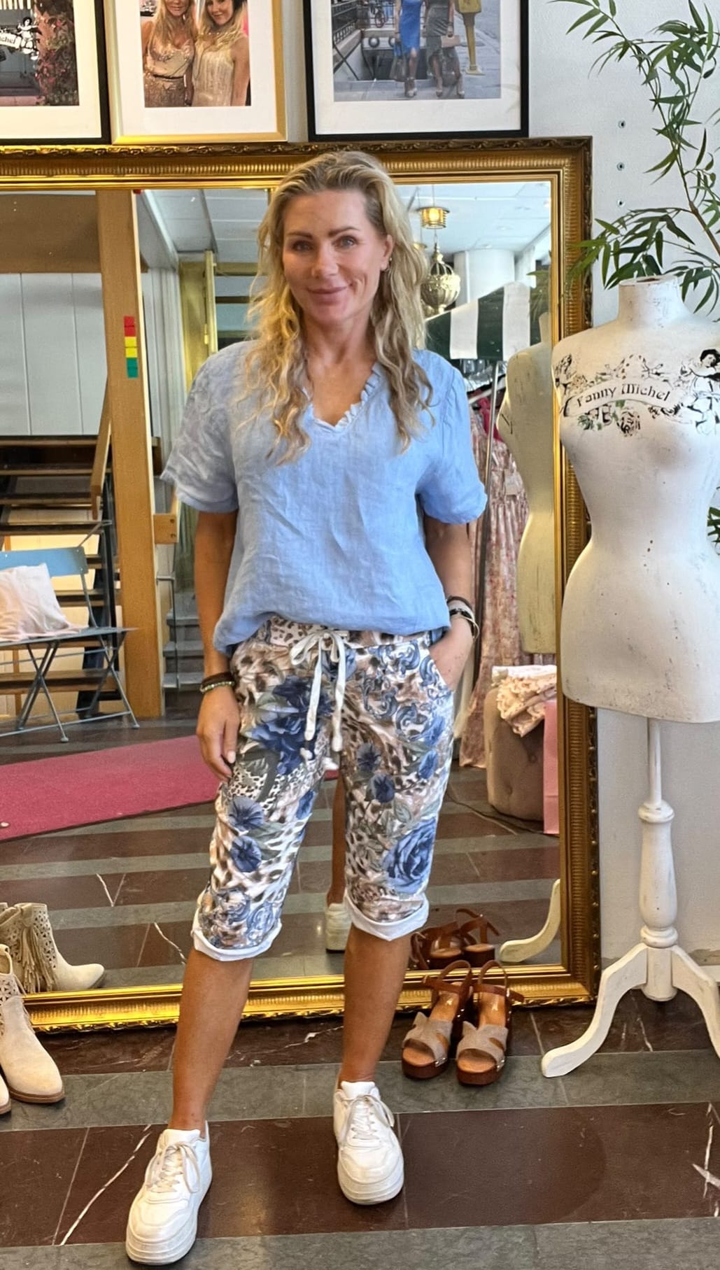 Kvinna står och poserar avslappnat med ena handen i fickan i en klädbutik, framför en stor guldramsspegel. Hon bär ljusblå blus, blommiga capribyxor och vita sneakers; provdocka bredvid med texten ”Fanny Michel”.