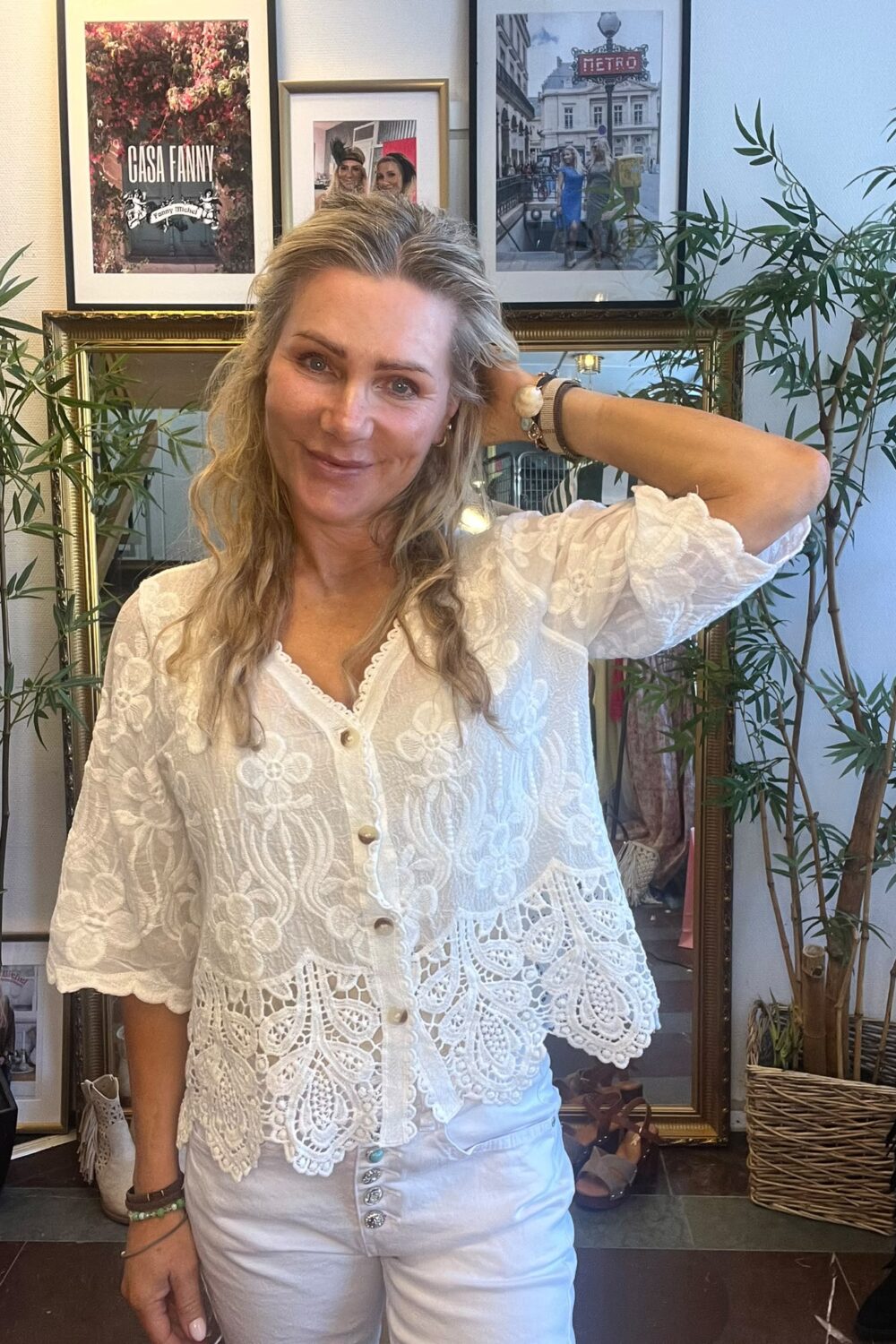 Leende kvinna i vit broderad blus och vita byxor poserar med ena handen bakom huvudet inomhus. Bakom henne syns en stor spegel, gröna krukväxter och inramade foton med texten “CASA FANNY” och “METRO”.