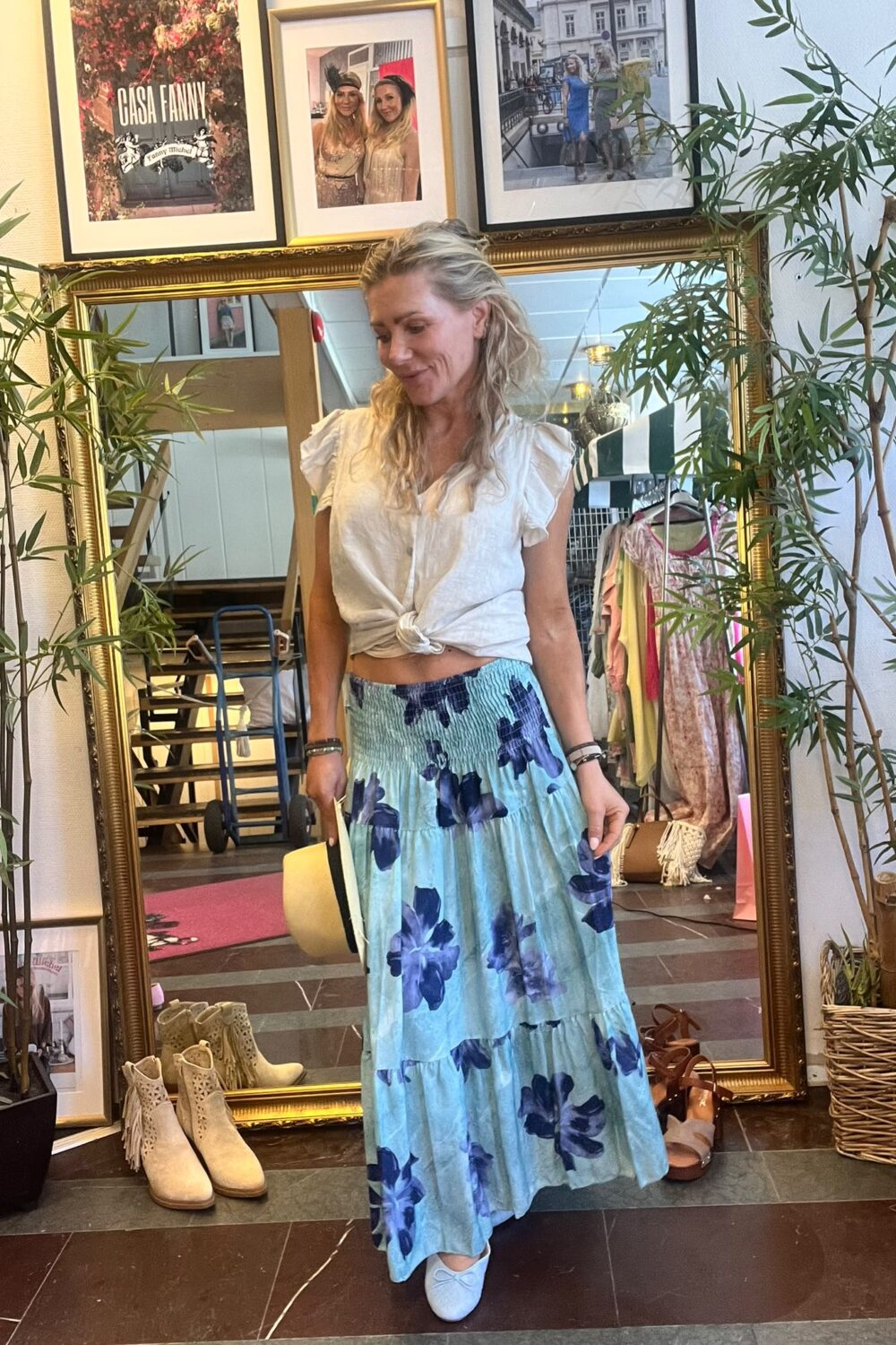 Kvinna står framför en stor guldspegel och provar outfit, håller en ljus hatt och ser ner med ett leende. Hon bär vit knuten blus och lång blåblommig kjol i en klädbutik med växter och skor. Text på väggbild: “CASA FANNY”.