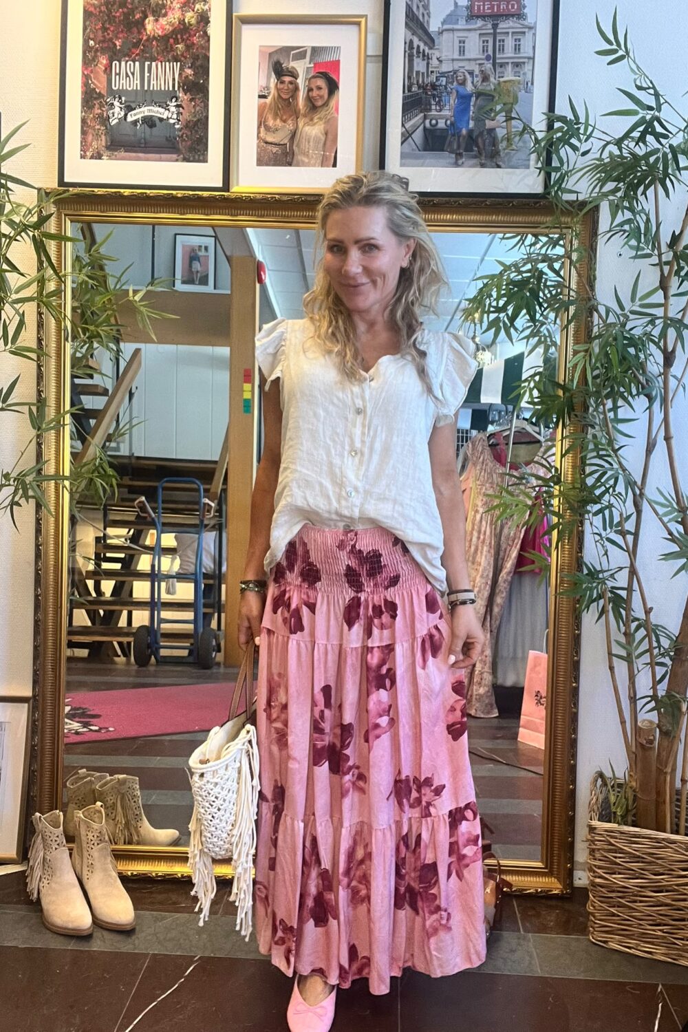 Kvinna står och ler framför en stor guldramad spegel i en butiksmiljö, klädd i vit blus och rosa blommig långkjol med rosa skor och vit fransväska. På väggen syns texten ”CASA FANNY”.