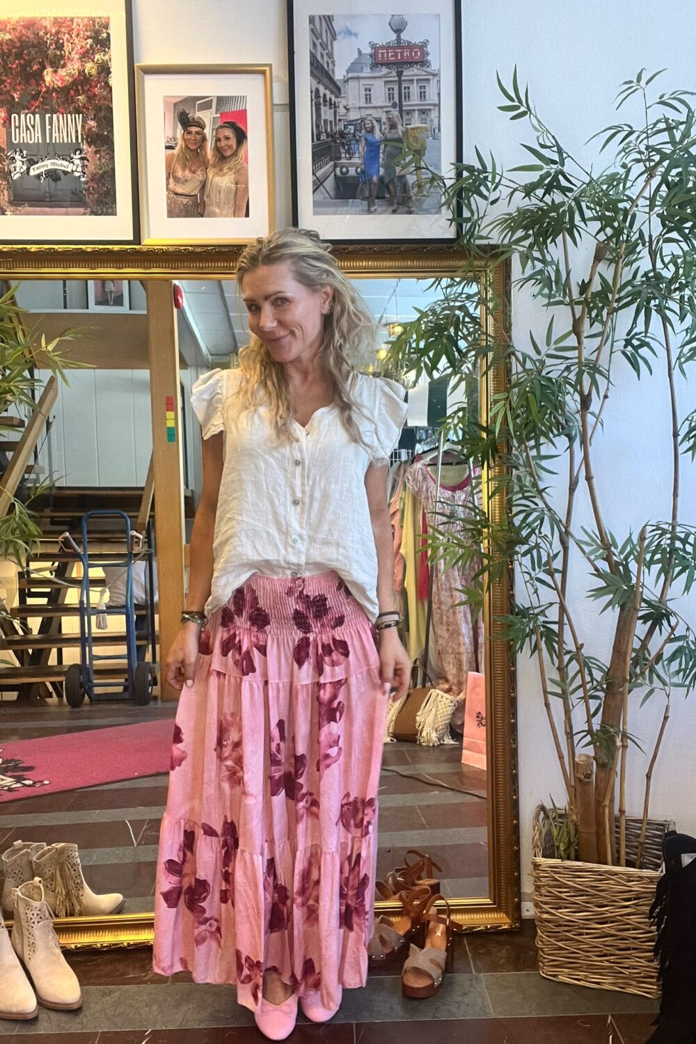 Blond kvinna står och ler framför en stor guldramsspegel i en klädbutik, klädd i vit blus och rosa blommig långkjol med rosa skor. Runtom syns skor, klädställning, krukväxt och tavlor med texten “CASH FANNY” och “METRO”.